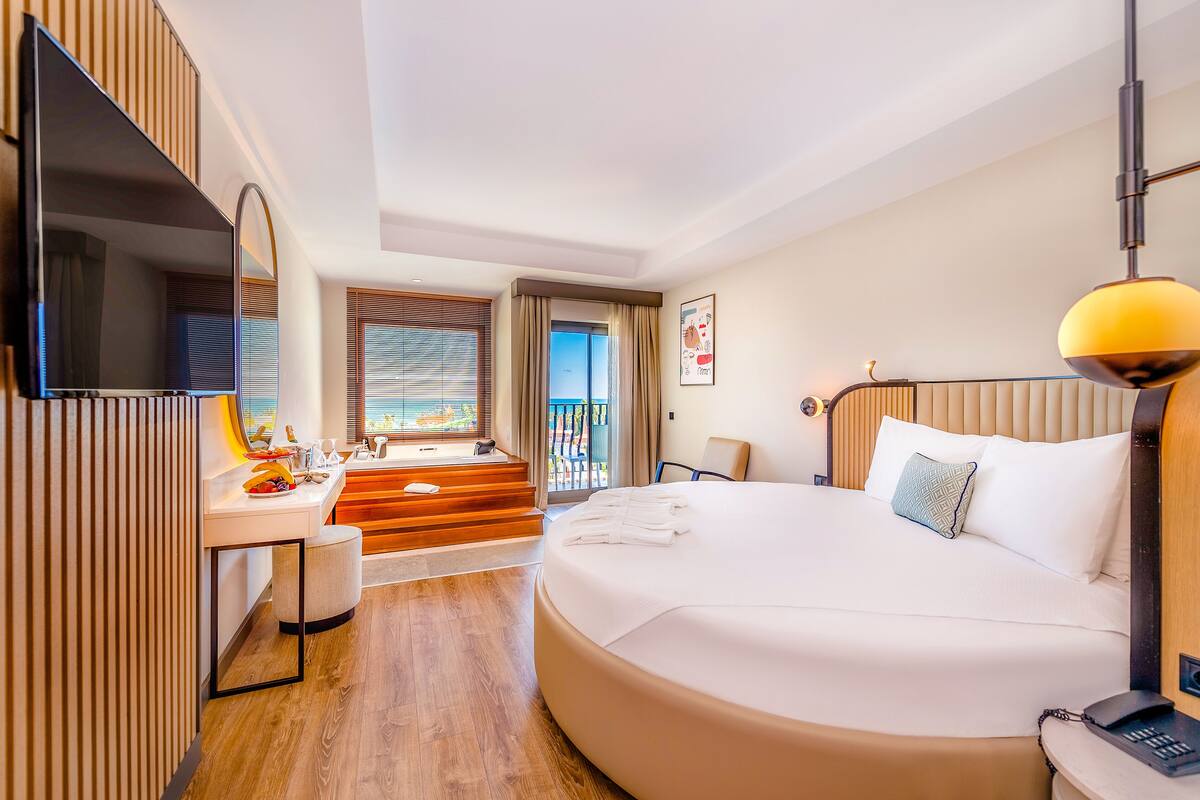 A hotel room with a large bed, a desk with a flat-screen TV, a bathroom with a shower, and a balcony with a view.