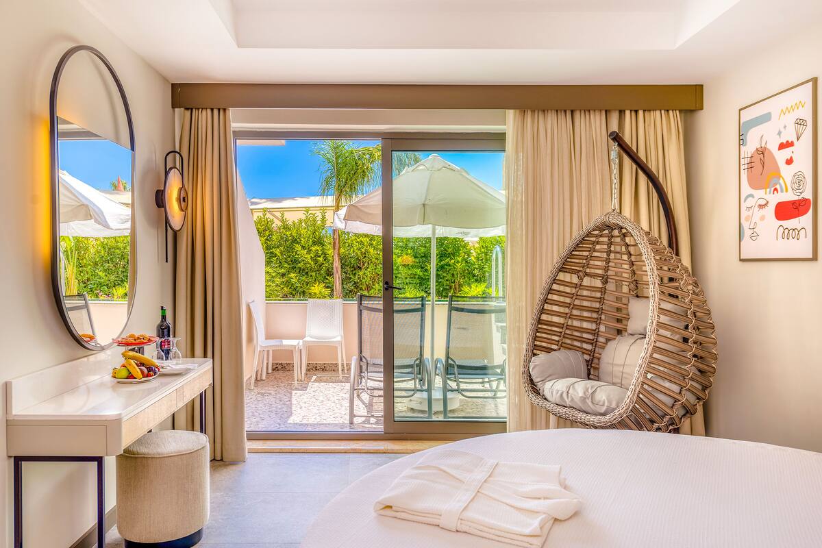 A hotel room with a bed, a hanging chair, a balcony with a view of greenery, and a dining area with a table and chairs.
