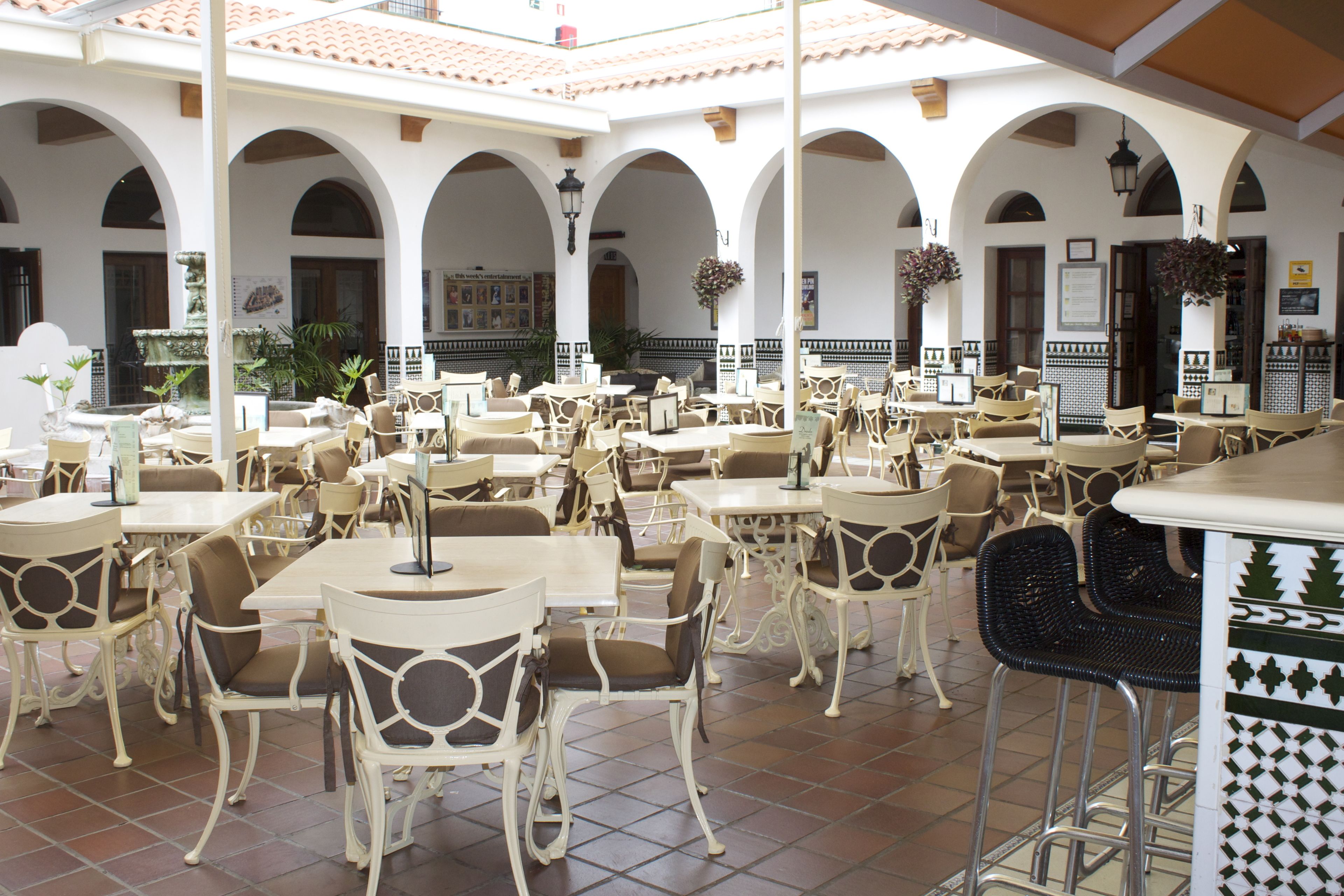 Un café extérieur spacieux, avec des tables et des chaises blanches, entouré d’arcades et agrémenté de plantes suspendues.
