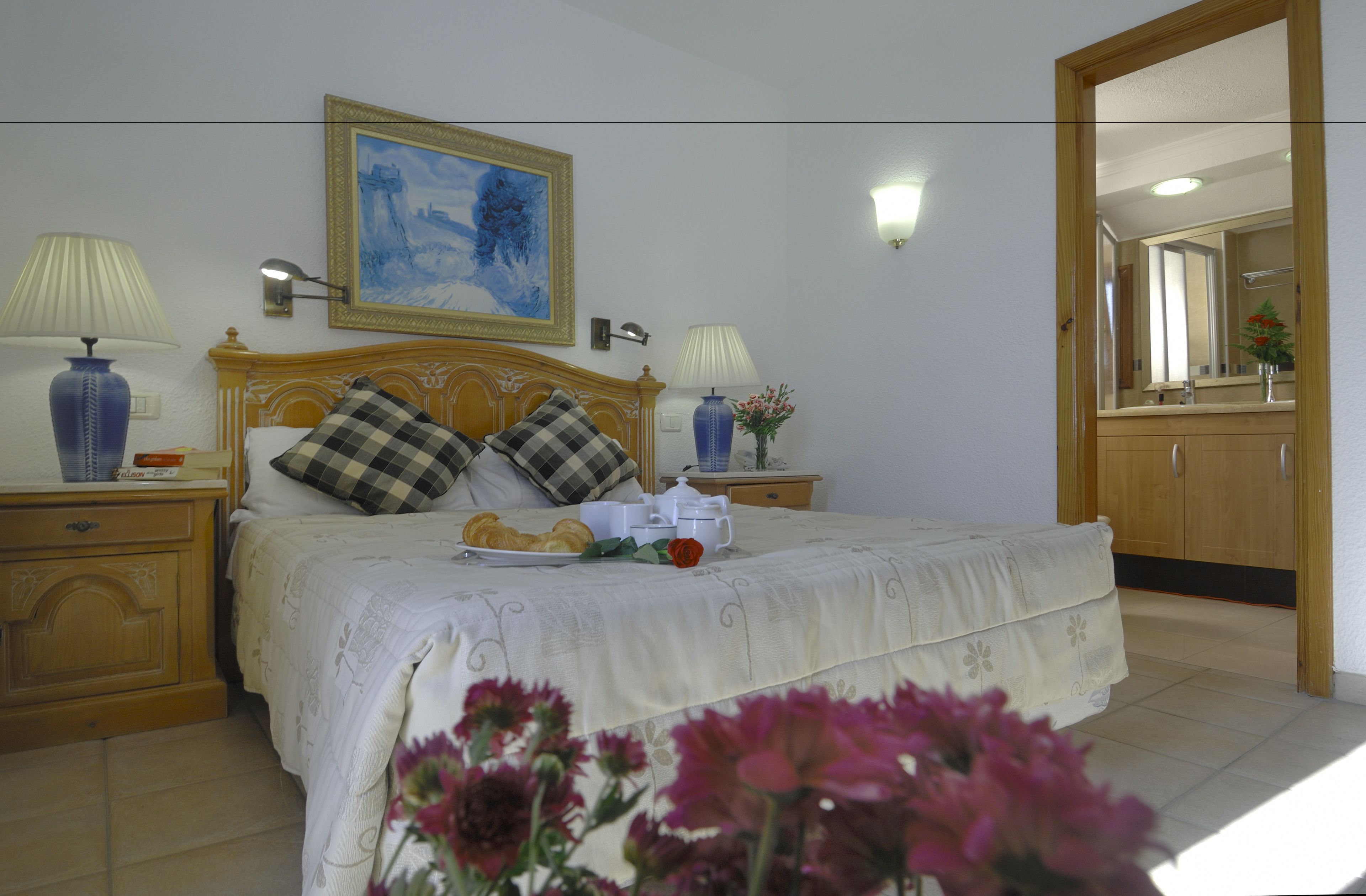 Un dormitorio con una cama, dos mesitas de noche, un tocador, un cuadro, un espejo y un baño visible a través de una puerta abierta.