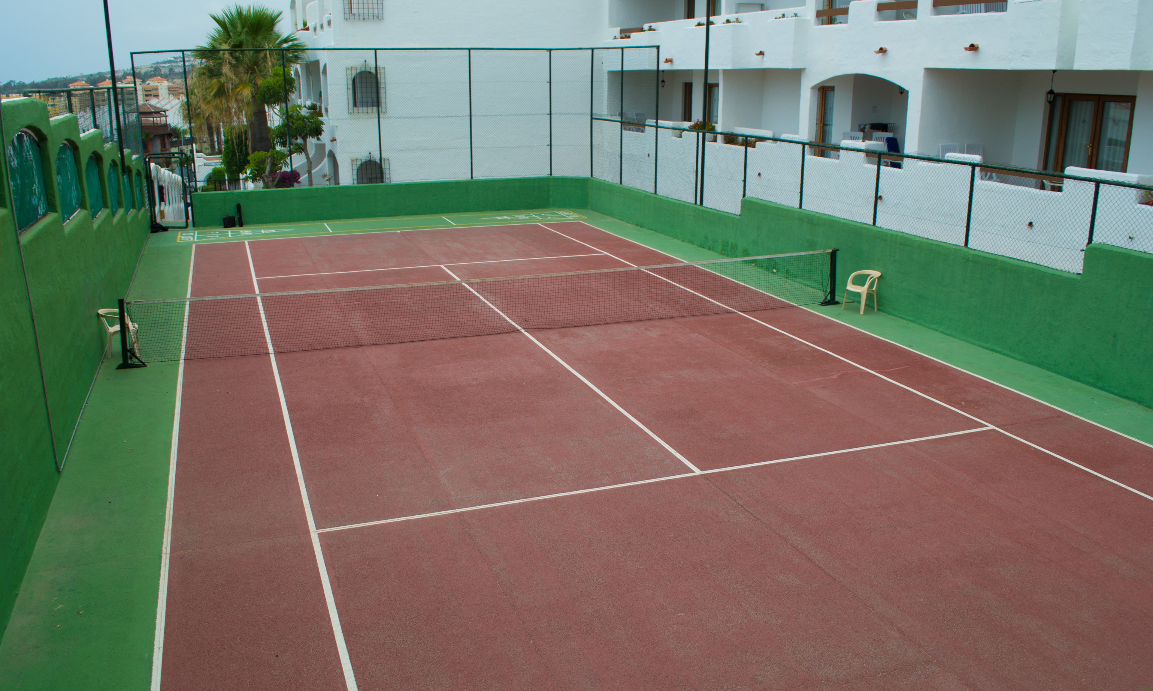 An indoor tennis court with a net, green and red flooring, and a white building in the background.