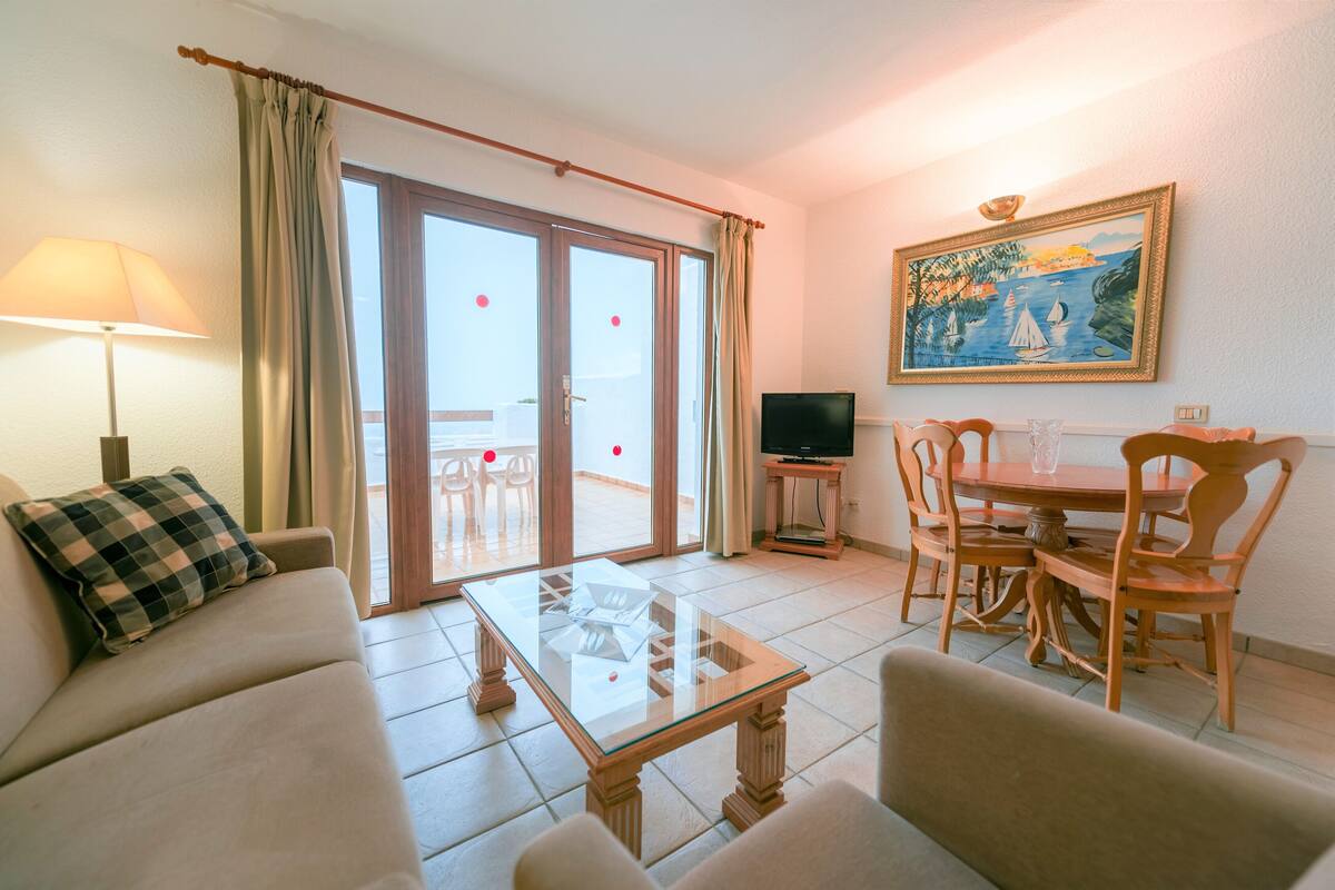 A living room with a sofa, a glass coffee table, a dining table with chairs, a TV, and a painting on the wall.