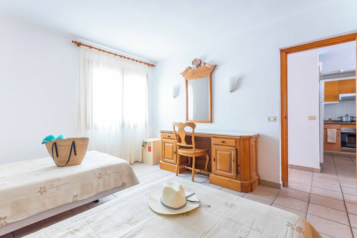 A room with a bed, a desk, a chair, a window with curtains, a mirror, and a view into the kitchen.