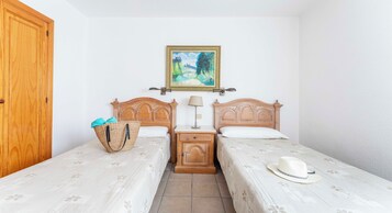 Una habitación con dos camas de madera, una mesita de noche con una lámpara, una cesta de mimbre y un sombrero sobre una de las camas.