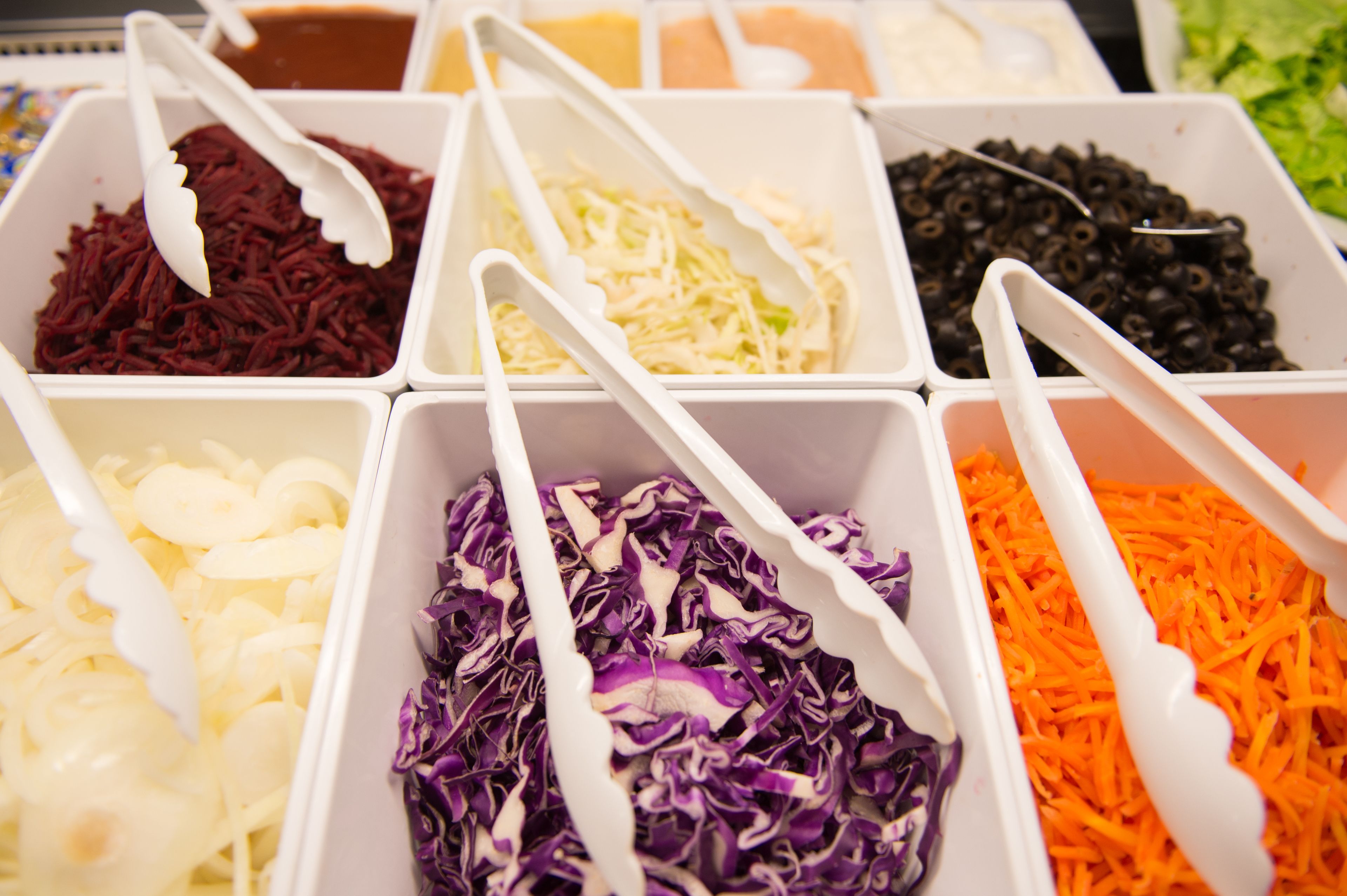 A variety of shredded vegetables in white containers with white plastic tongs.
