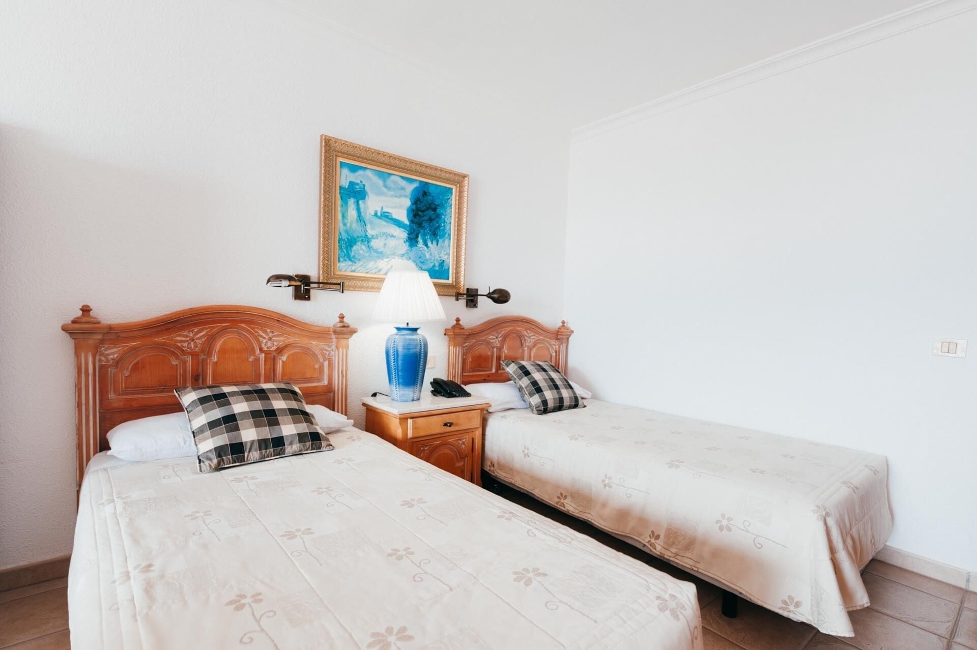 A hotel room with two beds, a nightstand with a lamp, a painting on the wall, and a telephone.