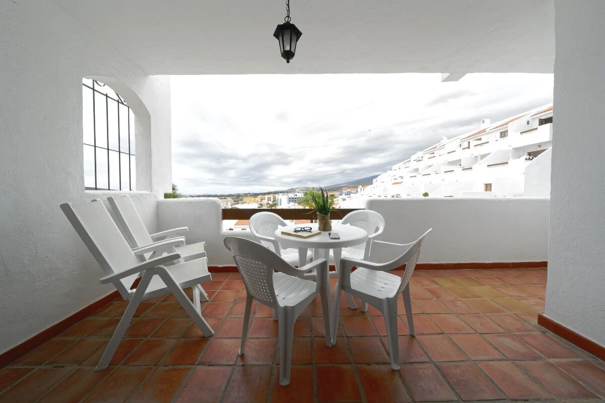 Een balkon met witte stoelen en een tafel, met uitzicht op een wit gebouw en een bewolkte lucht.