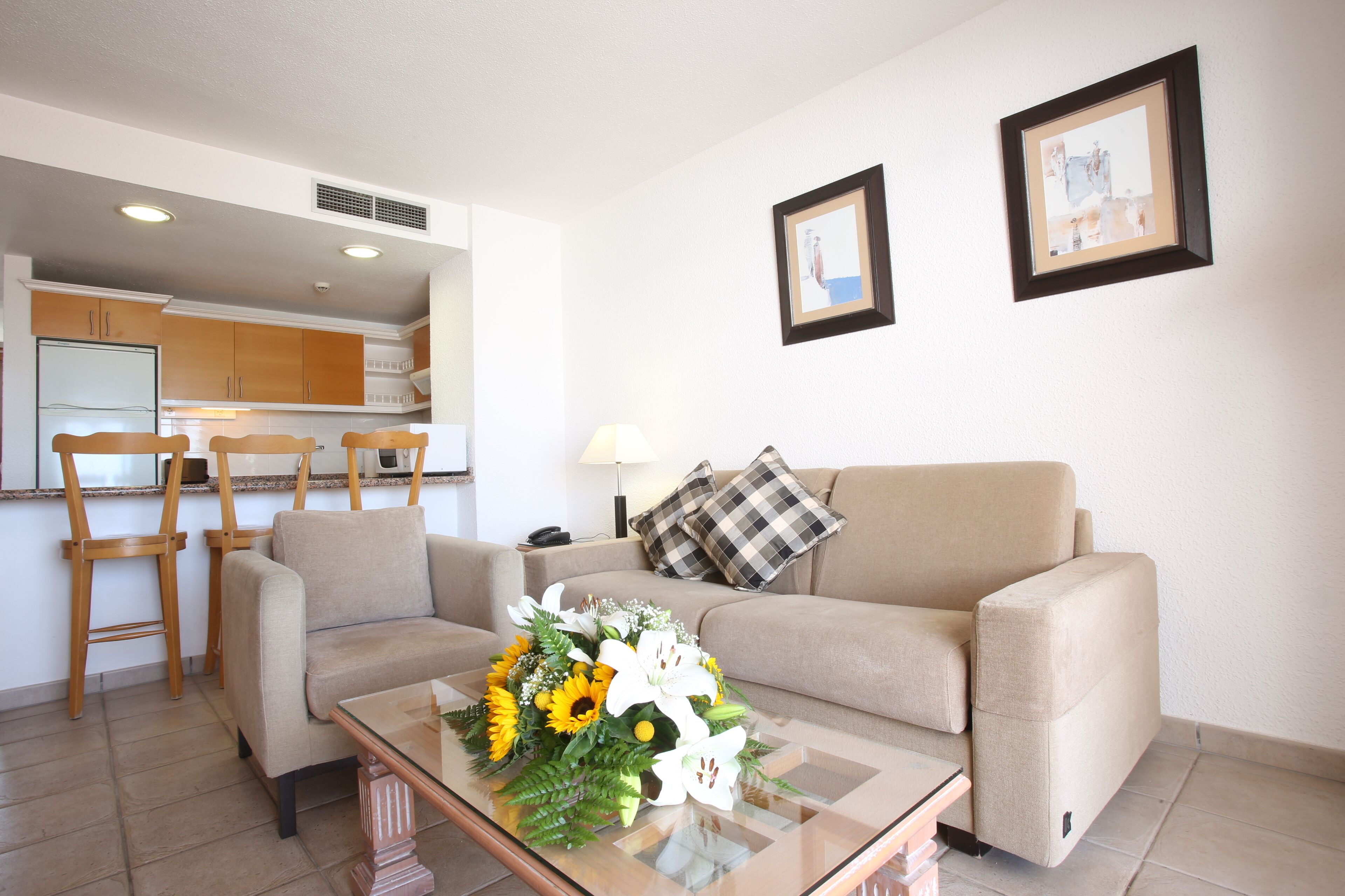 A living room with a sofa, a coffee table with a floral arrangement, a kitchen in the background, and two framed pictures on the wall.