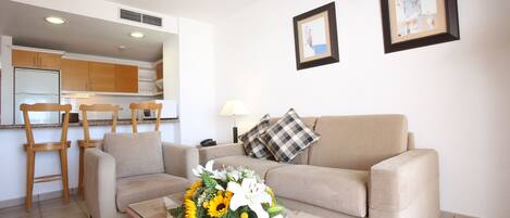 A living room with a sofa, a coffee table with a floral arrangement, a kitchen in the background, and two framed pictures on the wall.
