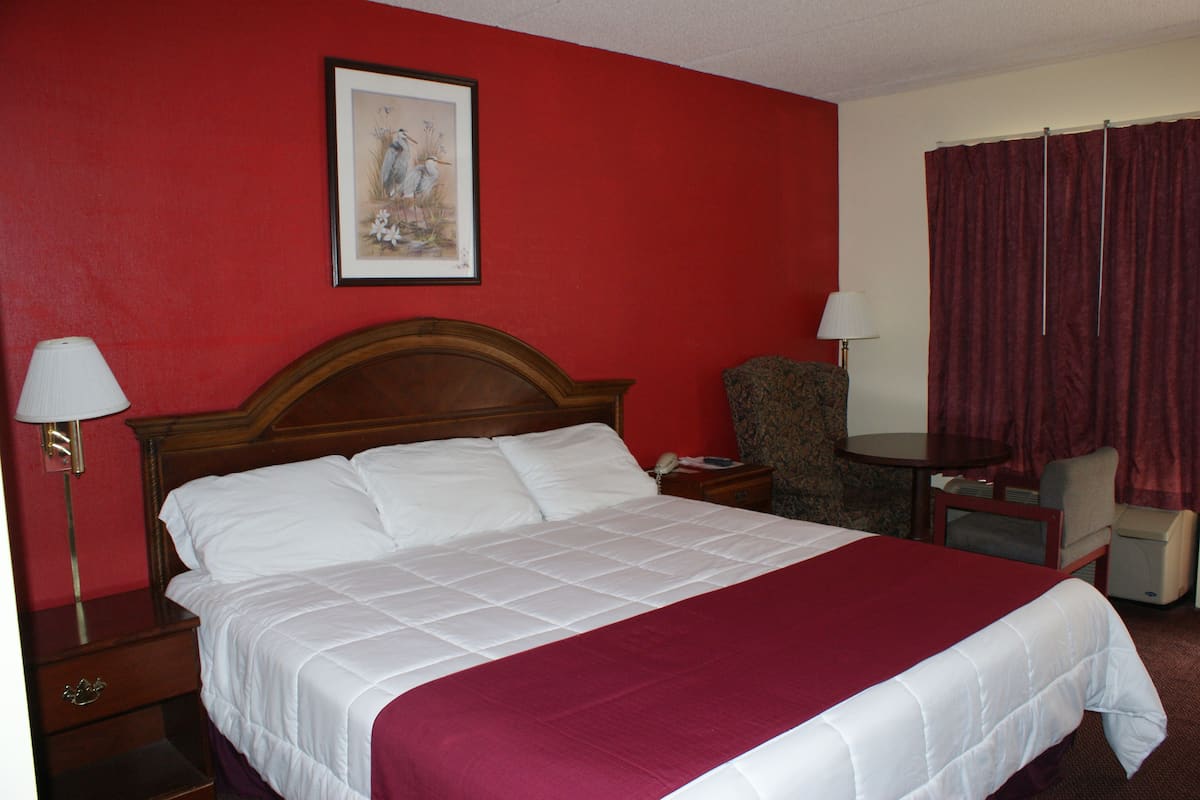 A hotel room with a large bed, a wooden headboard, two bedside tables with lamps, a chair, a small table, and a red curtain.