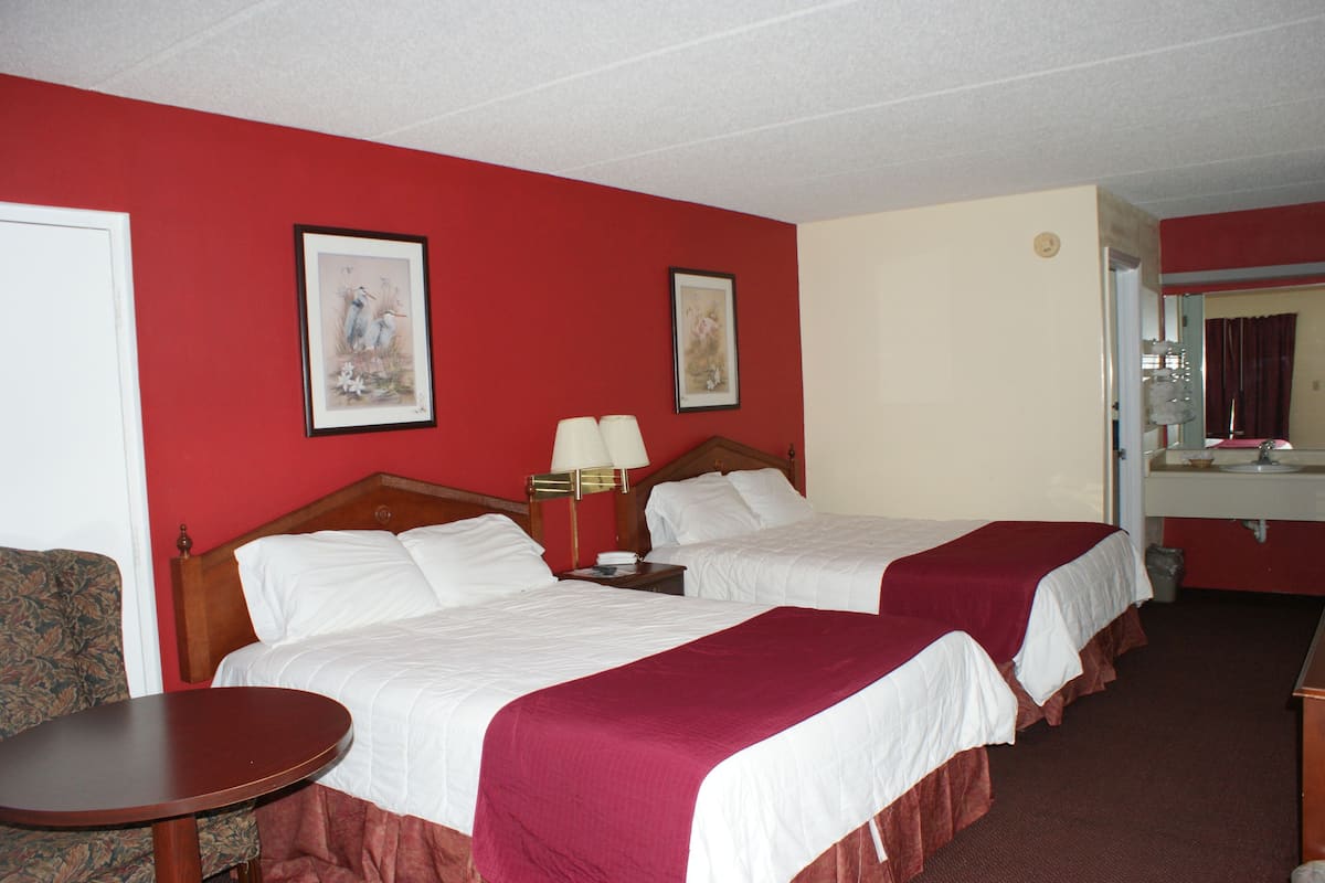 A hotel room with two beds, a small table, a chair, and a bathroom visible through an open door.