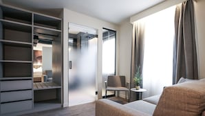 Una habitación de hotel moderna con armario empotrado, zona de estar con sofá y sillón, y una pequeña mesita con una planta.