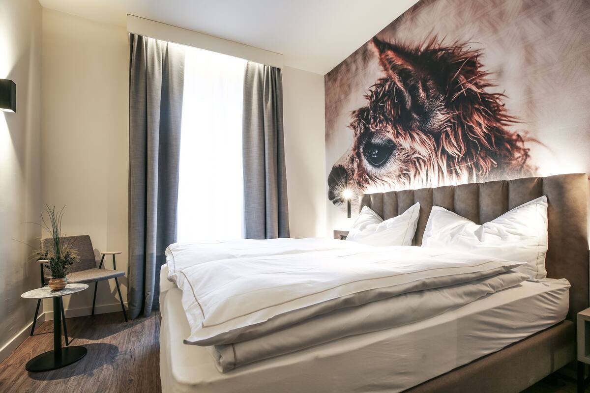A neatly made bed with white linens, a small round table with a plant, a chair, and a large wall art of a dog's face.