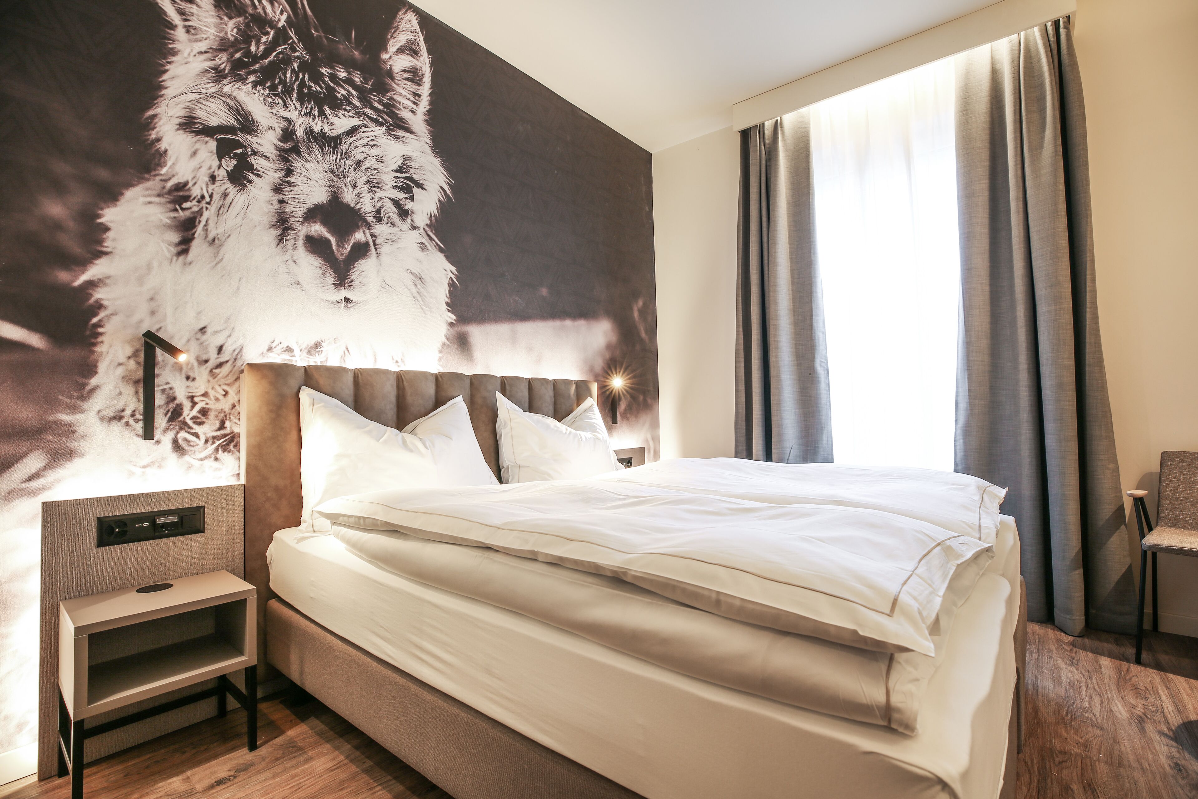A hotel room with a large wall mural of a llama, a bed with white linens, a bedside table, and a chair.