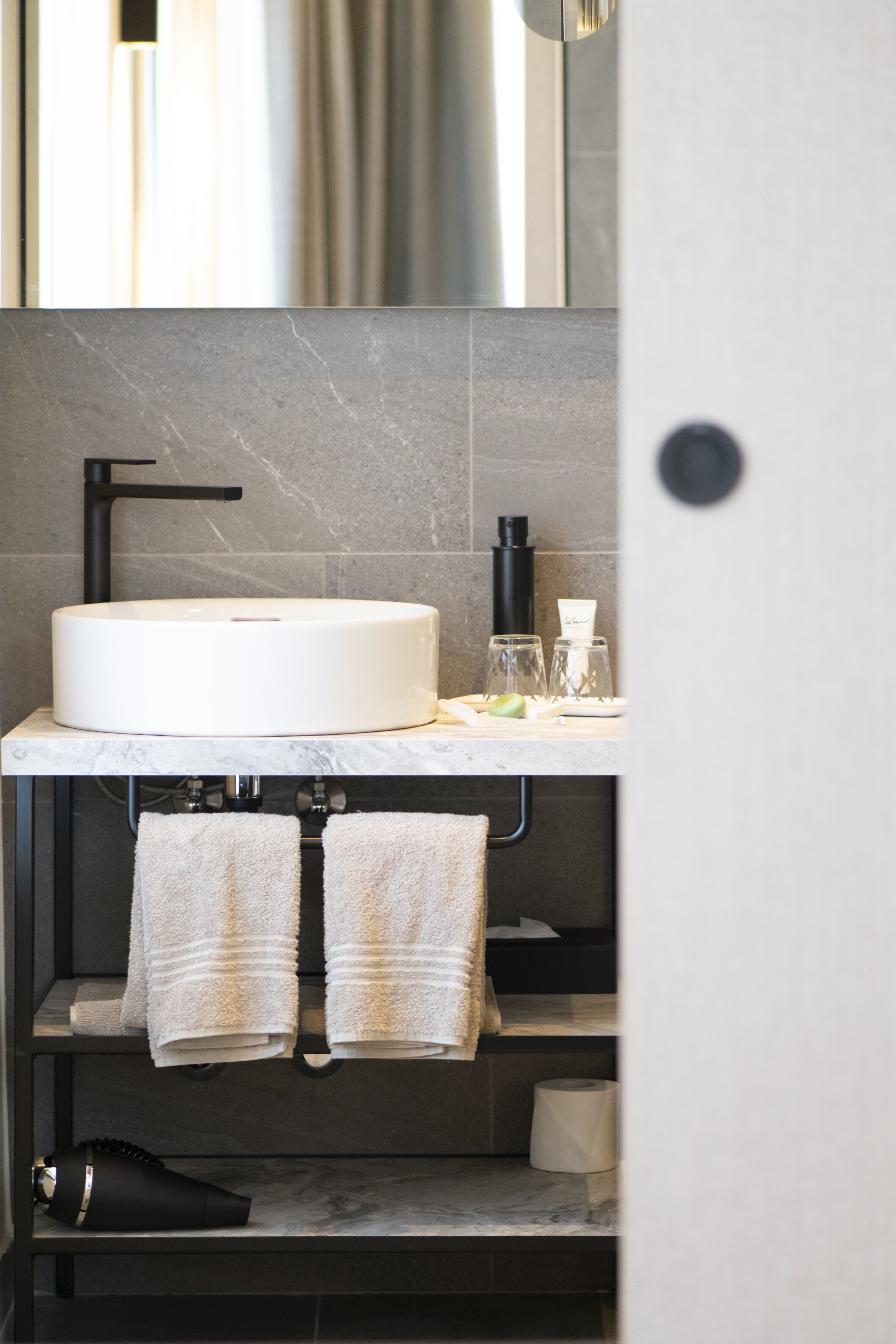 A modern bathroom with a round sink, black faucet, and marble countertop. There are two towels hanging on a rack, a black hairdryer, and a toilet paper roll on the shelf.