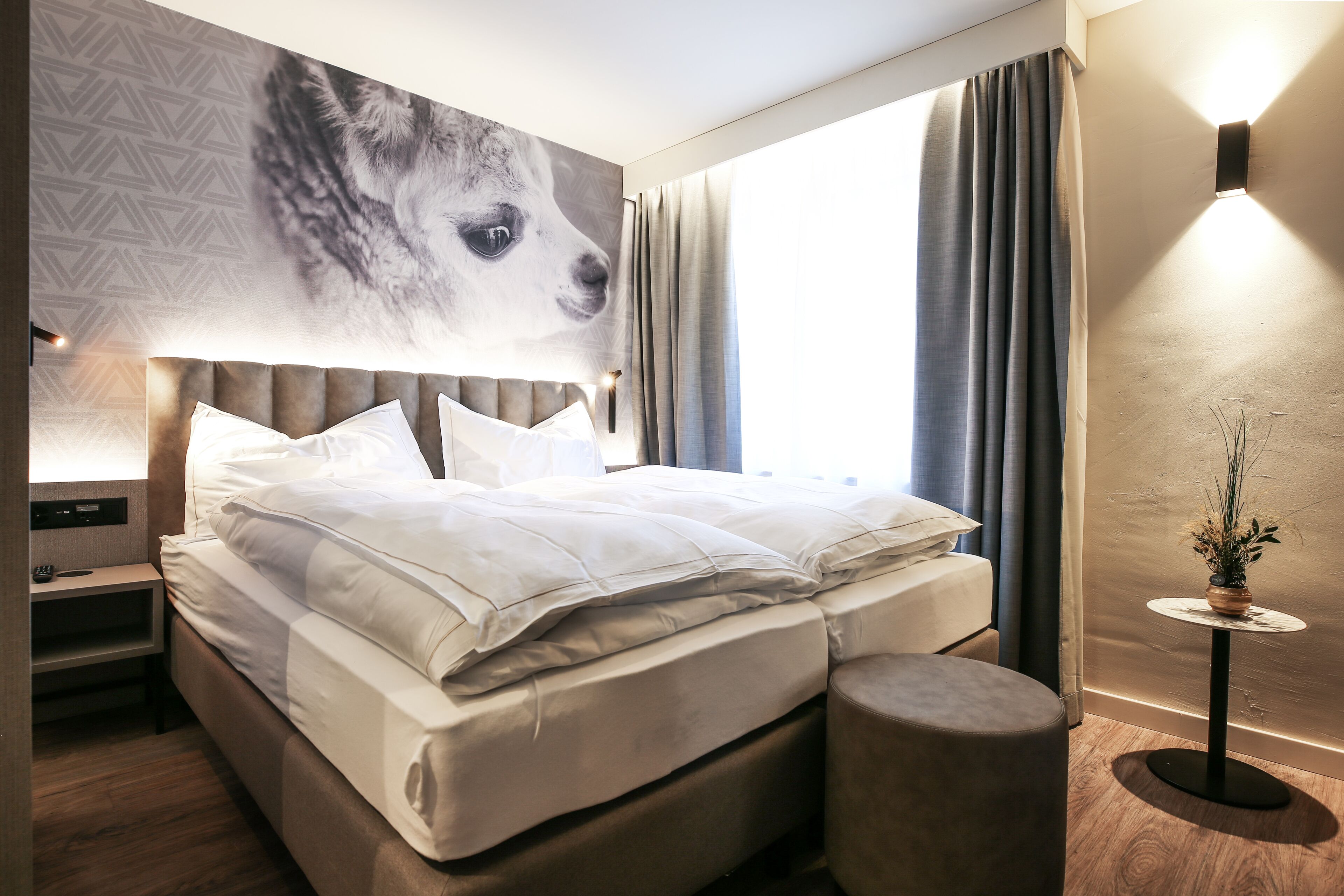 A modern hotel room with a large bed, a nightstand, a round side table, and a wall-mounted lamp.