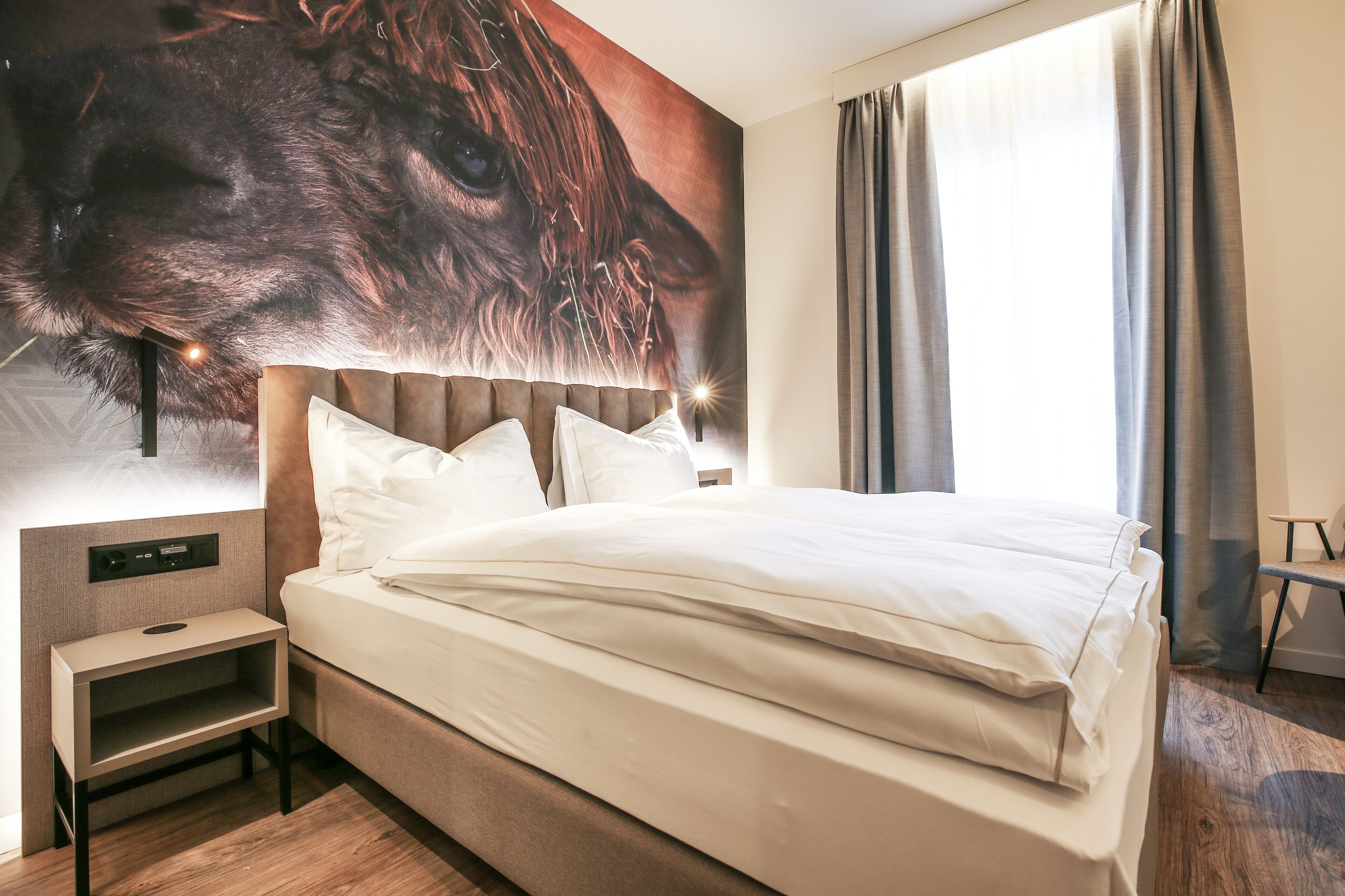 A neatly made bed with white linens, a headboard with a light wood finish, and a wall-mounted nightstand with a small shelf.