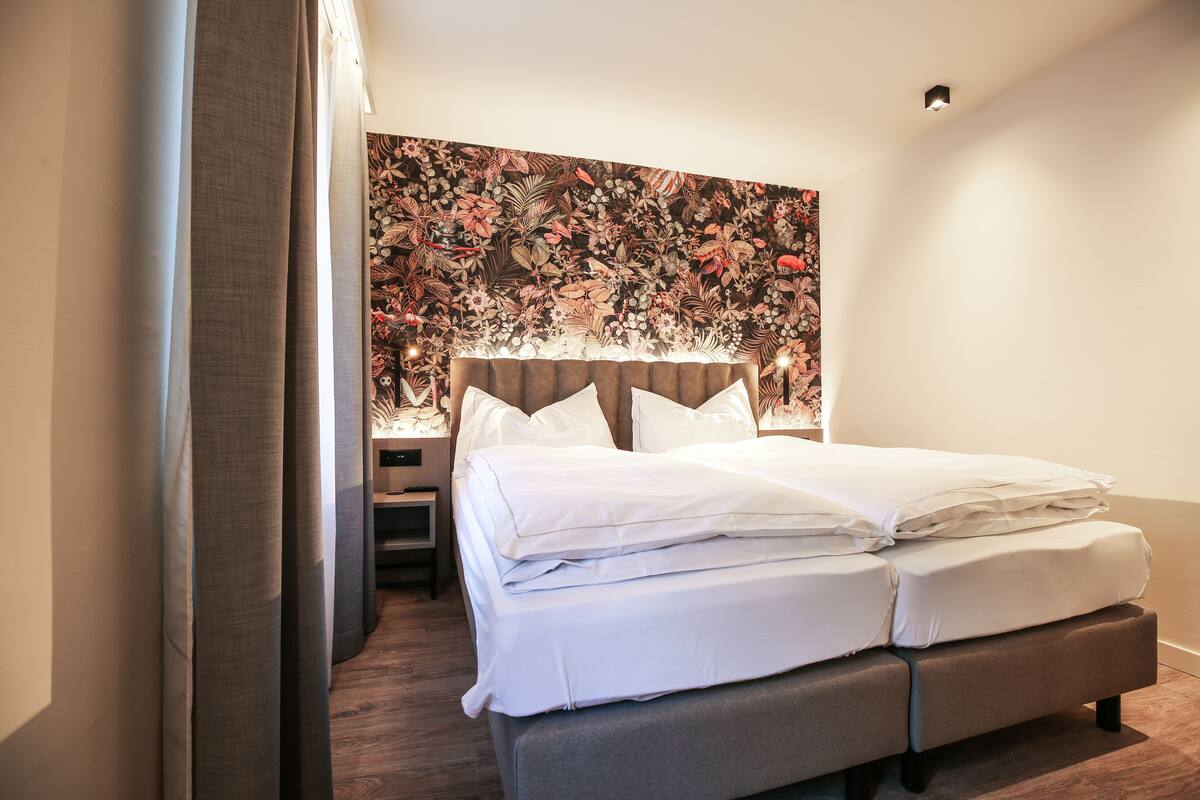 A neatly made bed with white linens, a headboard with a floral pattern, and a bedside table with a lamp.