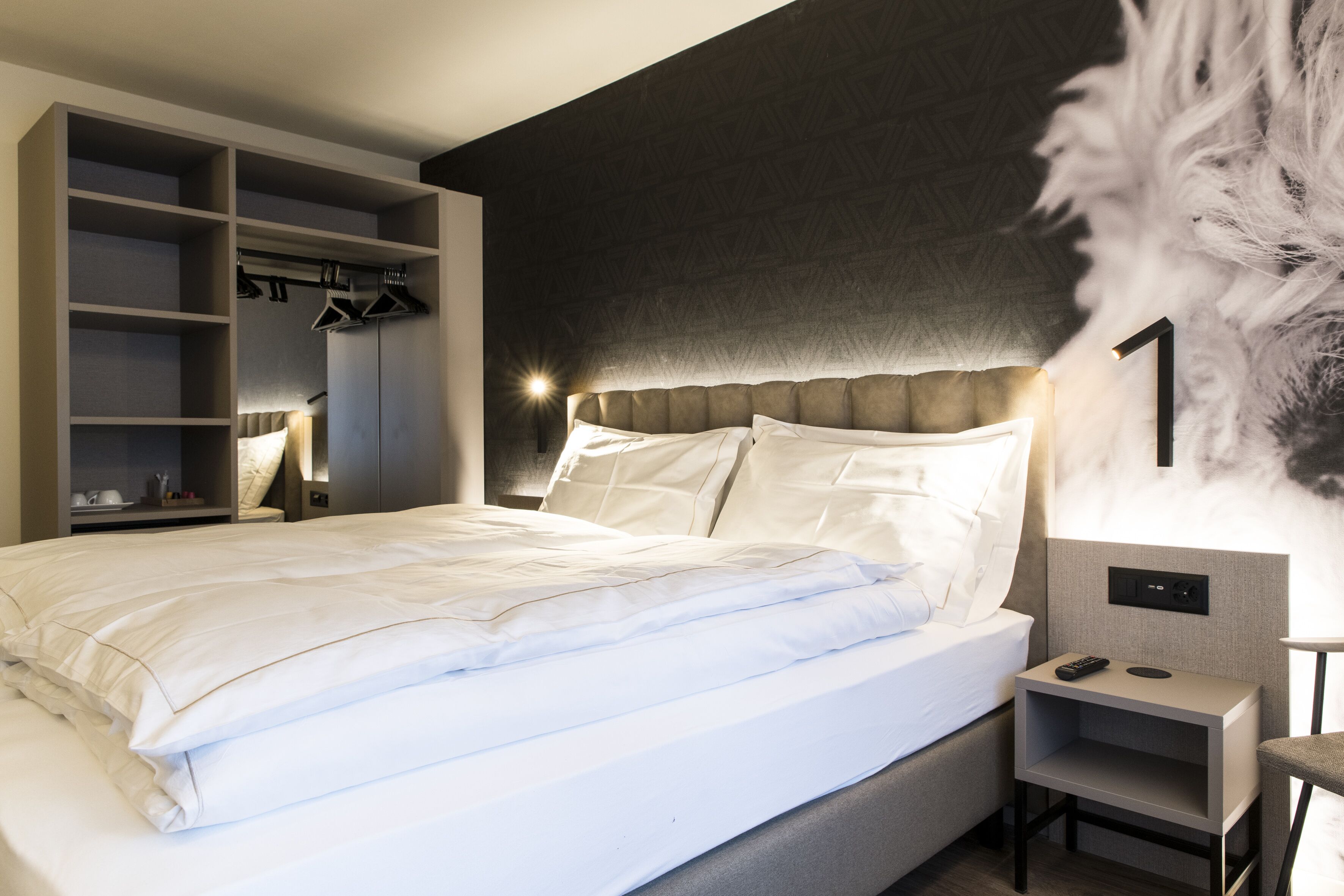 Quarto de hotel moderno com uma cama grande, guarda-roupa embutido, mesas de cabeceira e luminária fixa na parede.