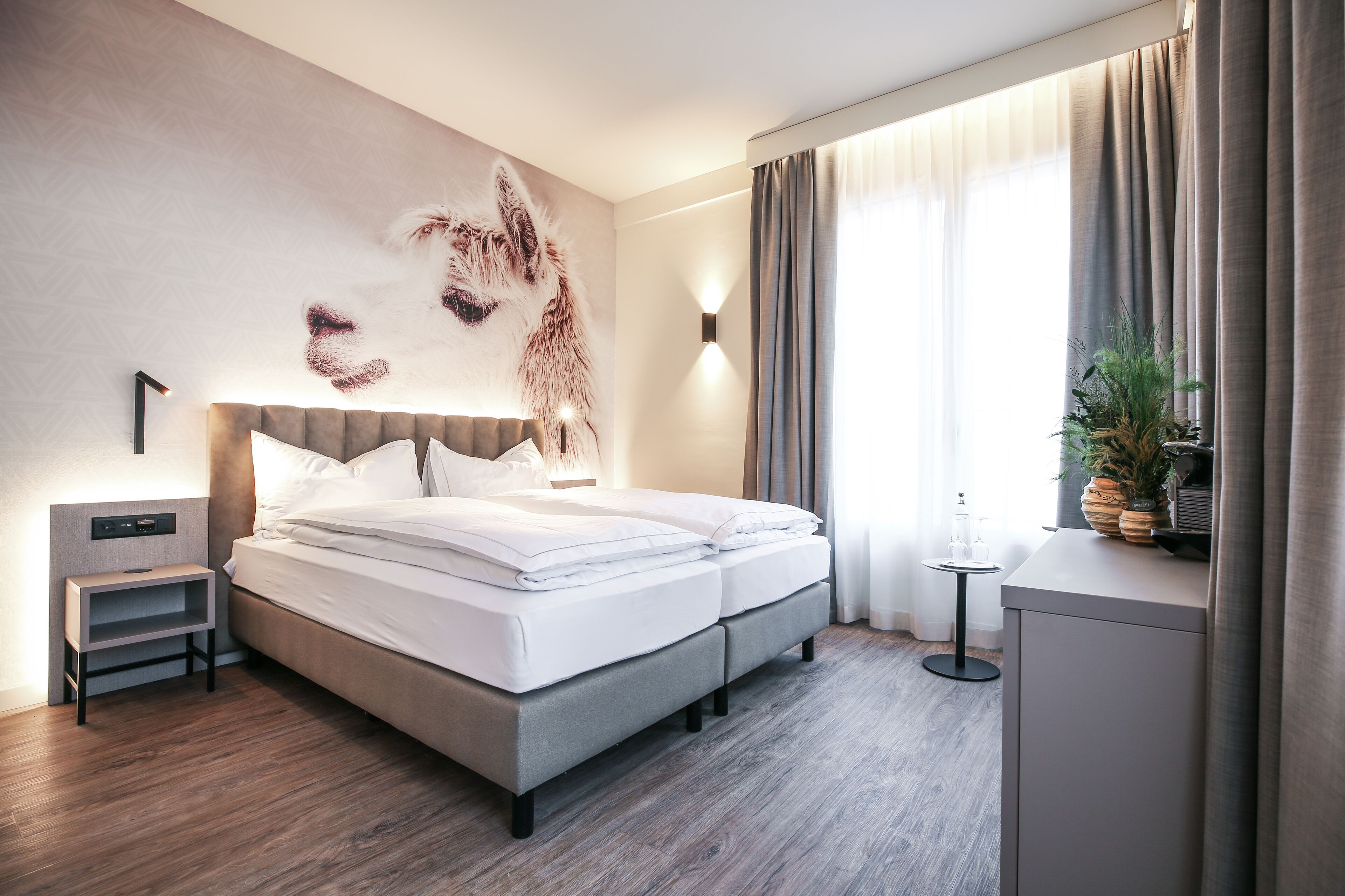 Quarto de hotel moderno com uma cama grande, uma mesa de cabeceira, uma luminária na parede e uma janela com cortinas.