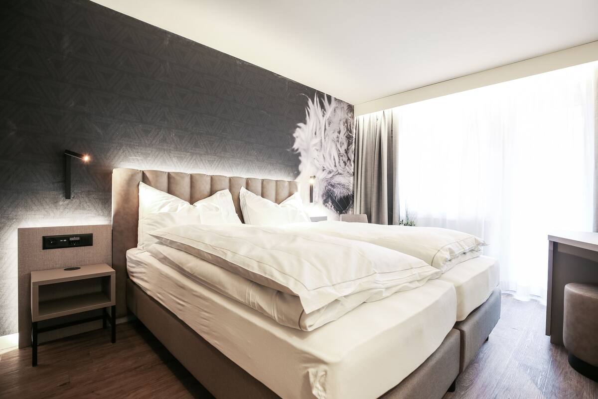 A modern hotel room with a large bed, a bedside table, a wall-mounted control panel, and a large window with curtains.