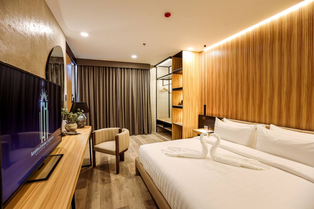 Habitación de hotel con una amplia televisión de pantalla plana, un sillón cómodo, una cama con ropa blanca y una toalla con forma de cisne, paneles de madera y un espejo.