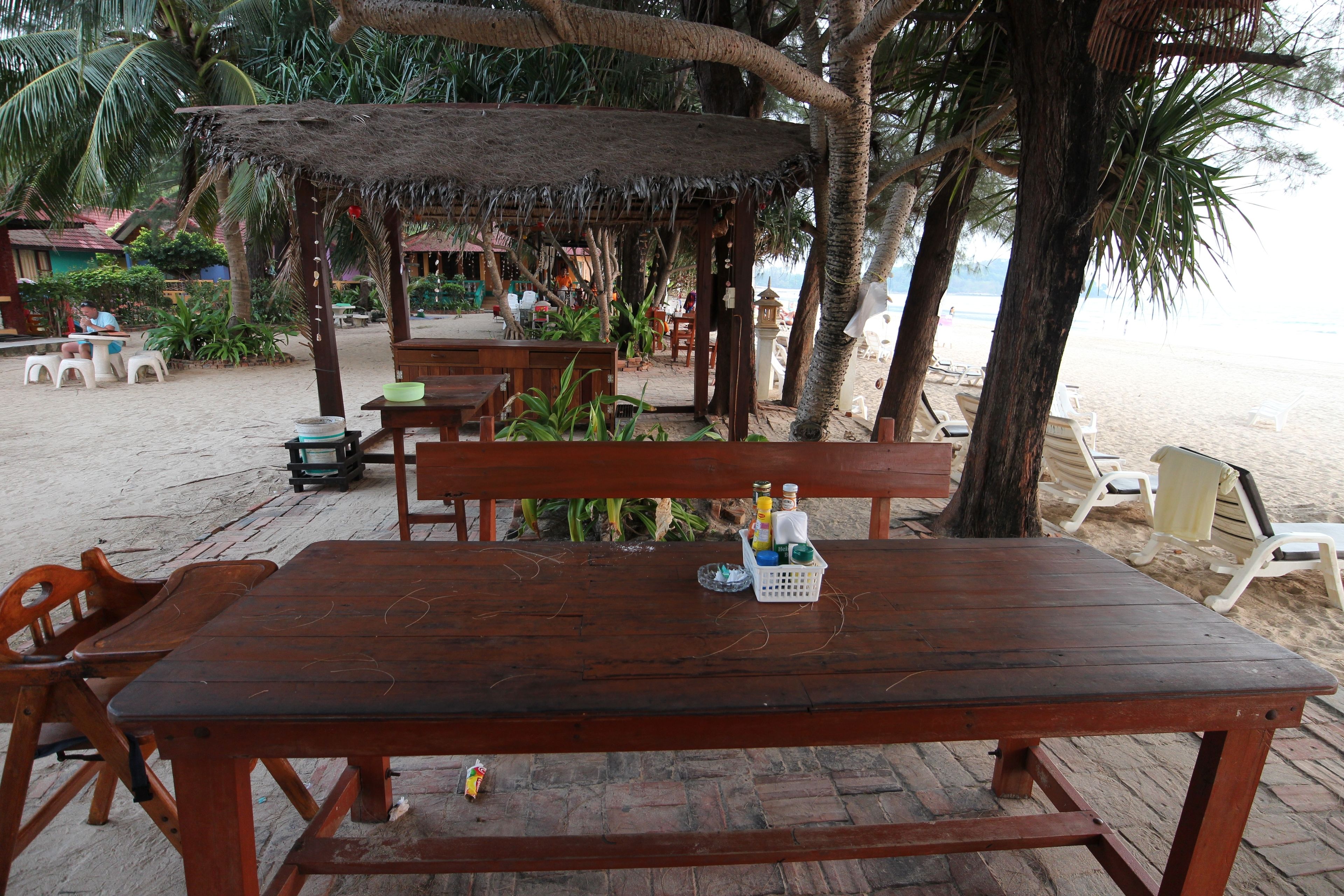 A wooden table with a basket of condiments, surrounded by chairs, under a thatched roof structure.