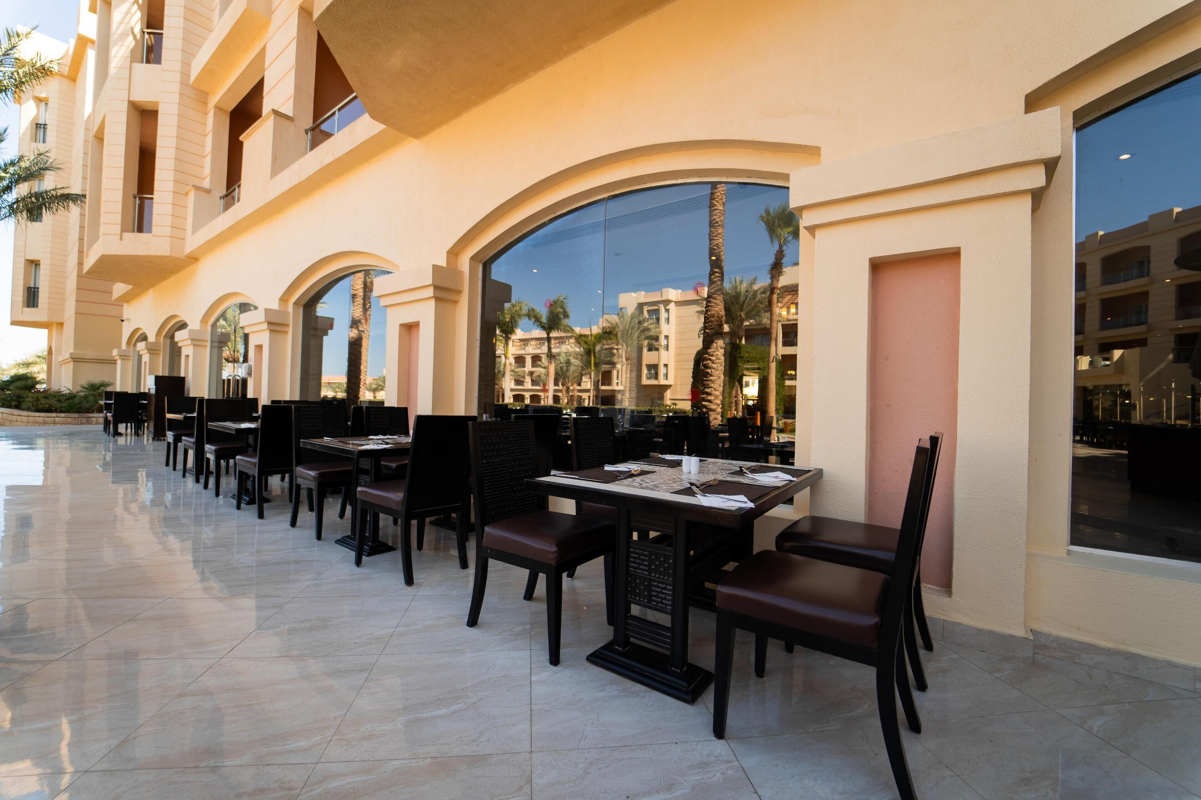 A row of outdoor dining tables with chairs, large windows, and a pool in the background.