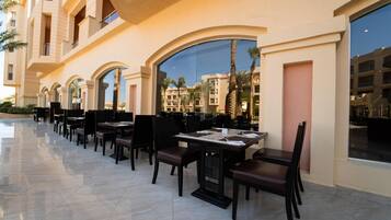 A row of outdoor dining tables with chairs, large windows, and a pool in the background.
