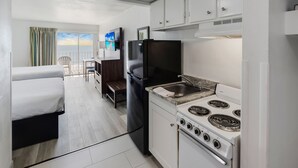 A compact kitchen with a refrigerator, stove, and sink. A bed and a small table are visible in the adjacent room.