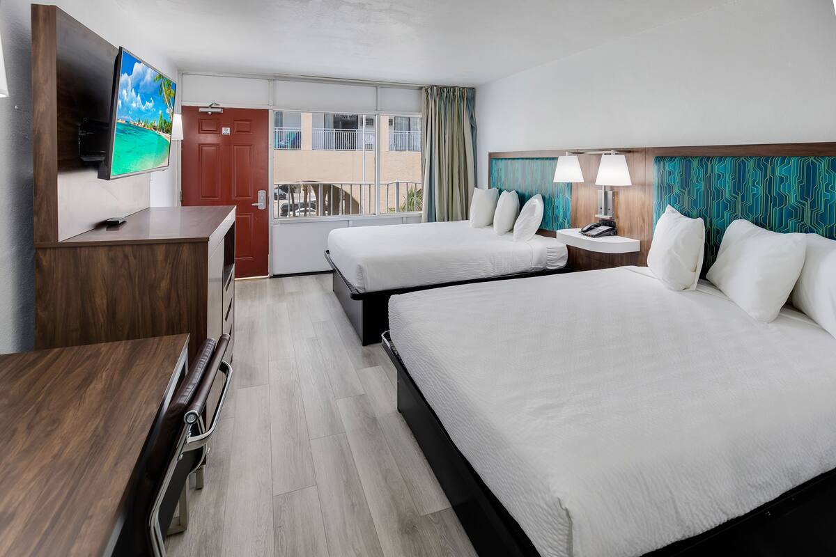 A hotel room with two beds, a TV, a desk, and a balcony with a view of a building.