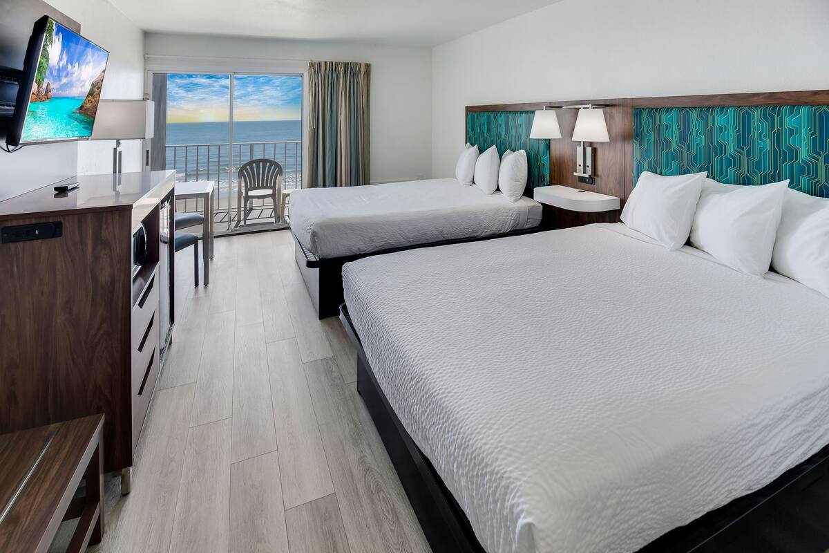 A hotel room with two beds, a desk, a chair, a TV, and a balcony with a view of the sea.