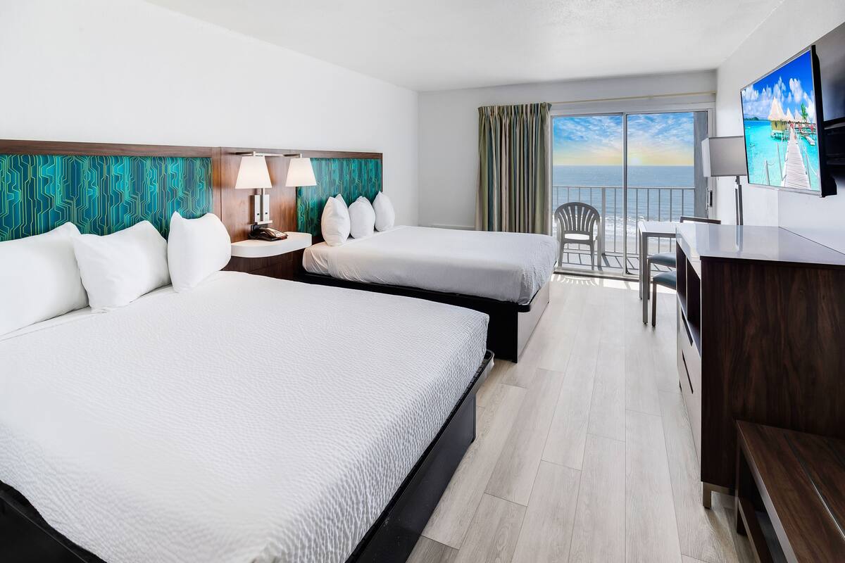 A hotel room with two beds, a desk, a TV, and a balcony with a view of the ocean.