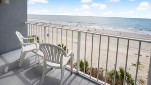 A balcony with white plastic chairs overlooking a beach with people and palm trees.