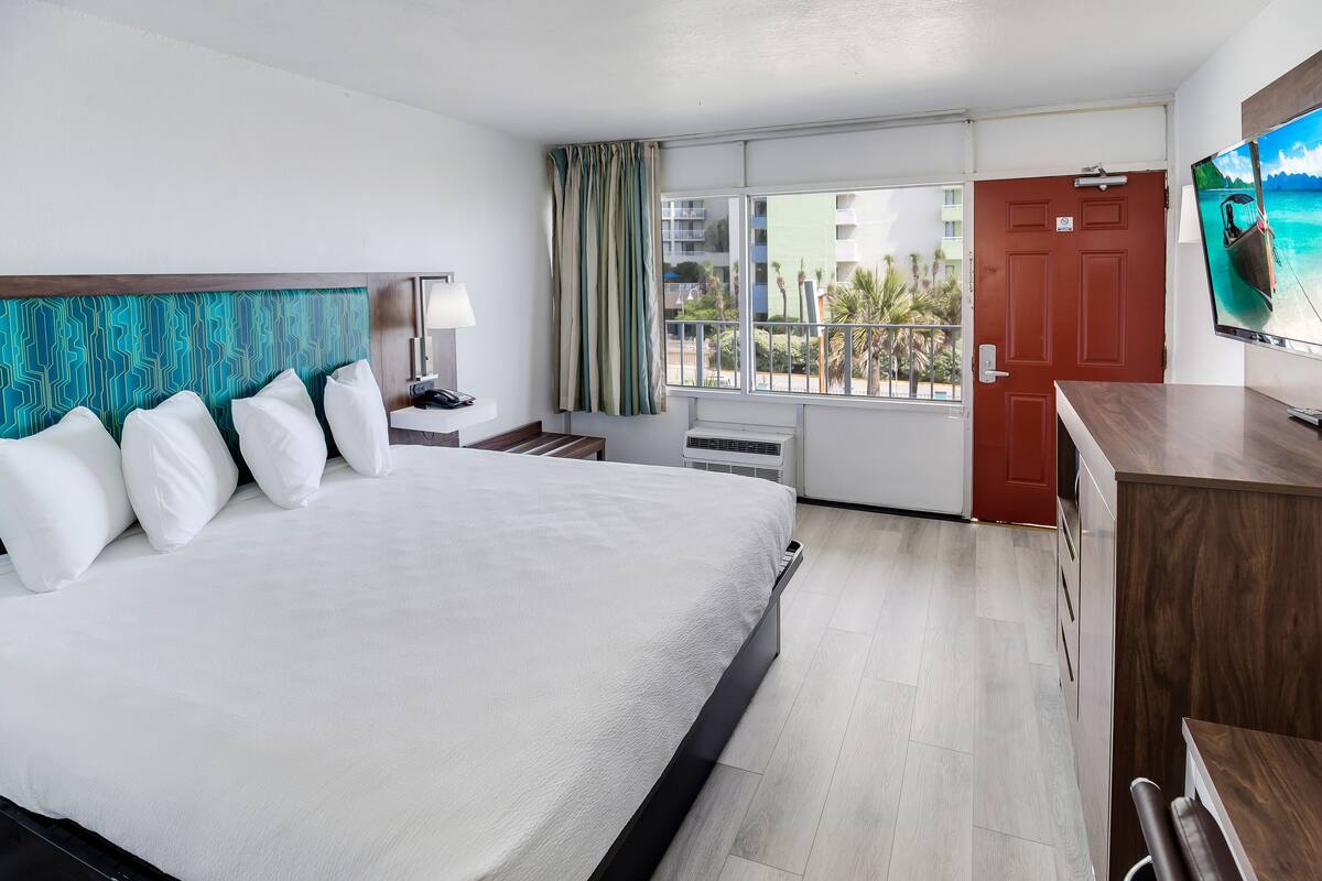 A hotel room with a large bed, a TV, a desk, a window with a view of buildings, and a red door.