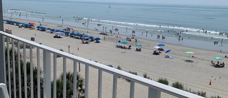 A beach view from a balcony with white railings, a sandy beach, and umbrellas.