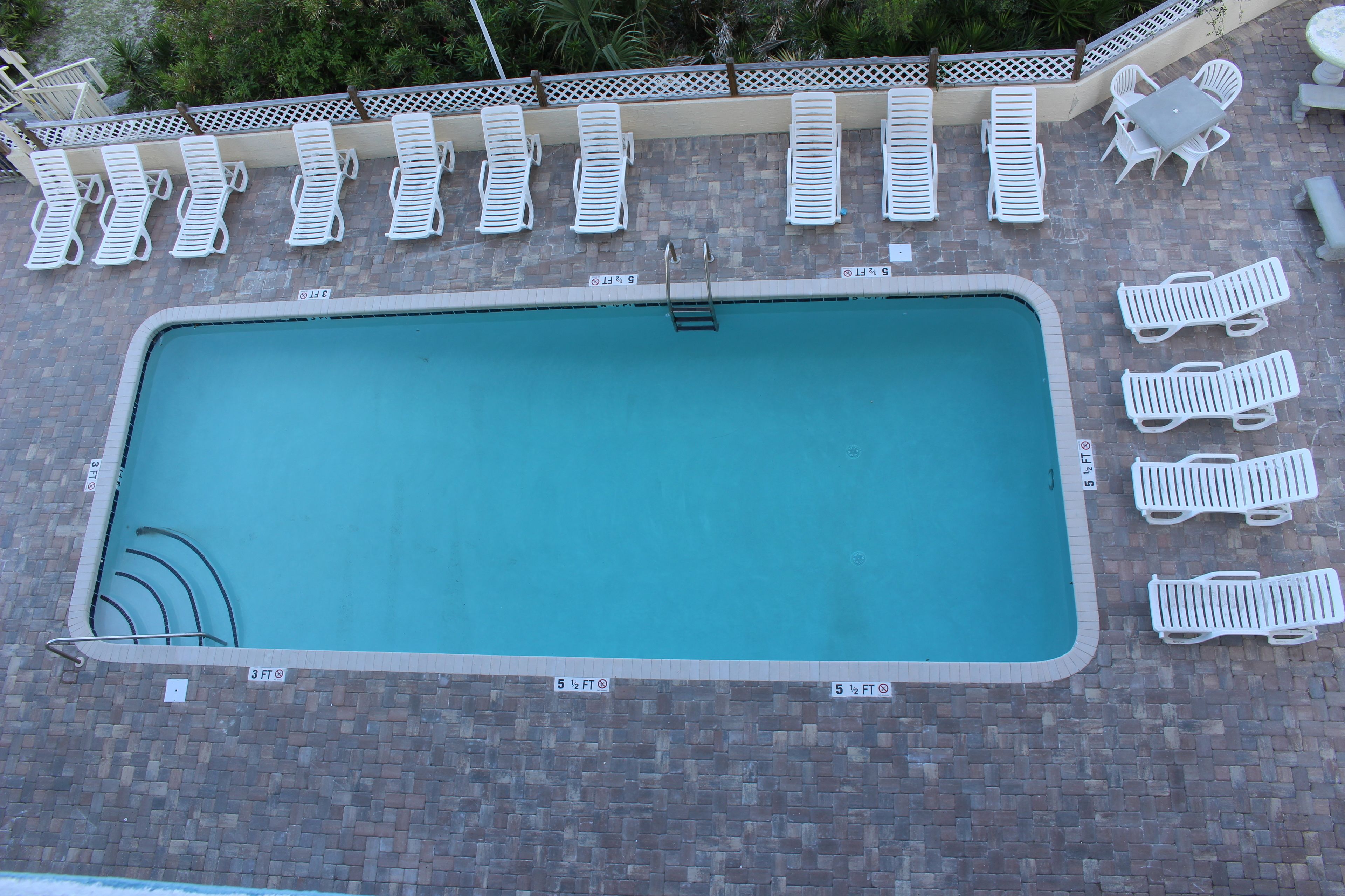A rectangular swimming pool with a brick border, surrounded by white lounge chairs and a small table with chairs.