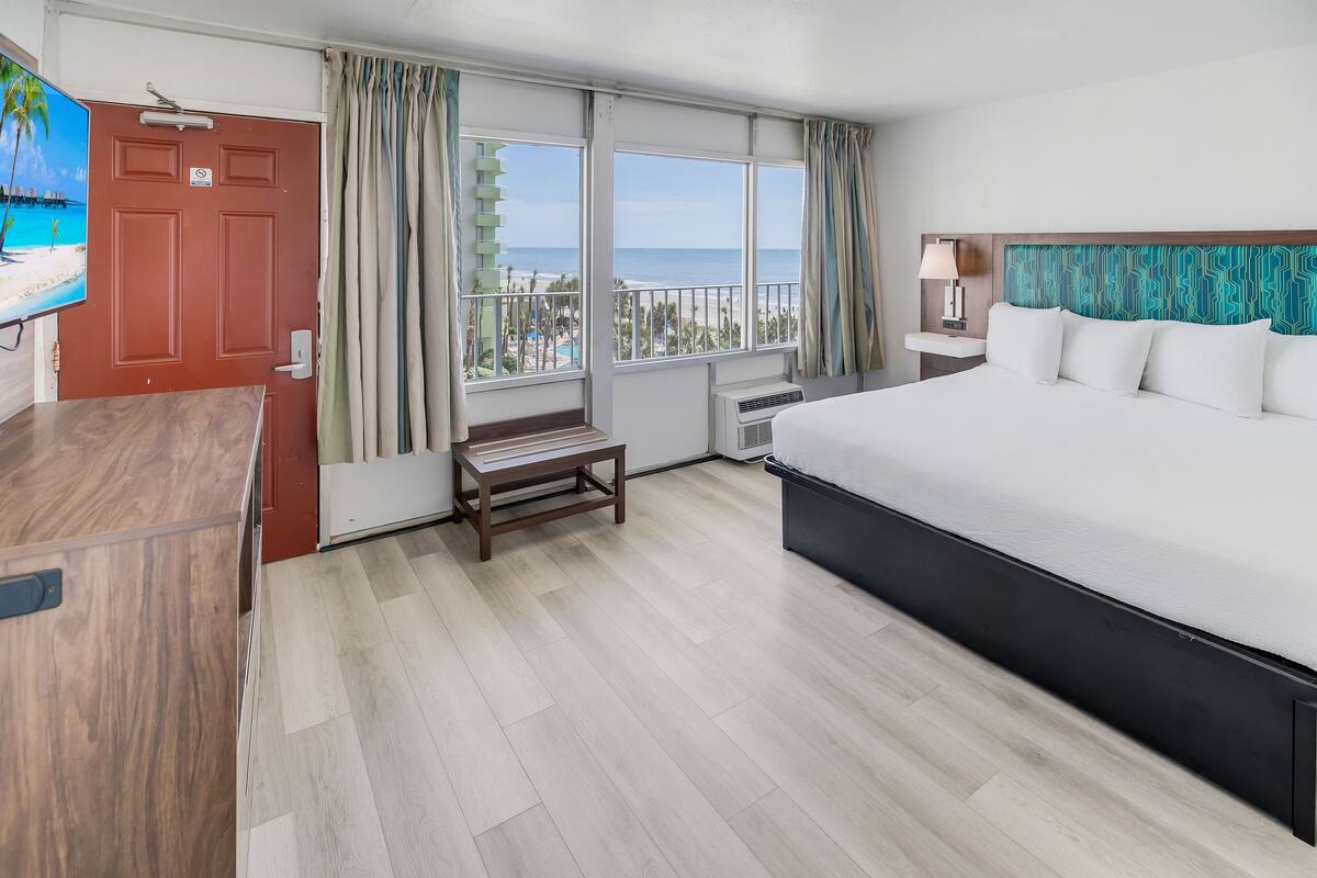 A hotel room with a bed, a desk, a TV, a balcony with a view of the beach and ocean, and a window with curtains.