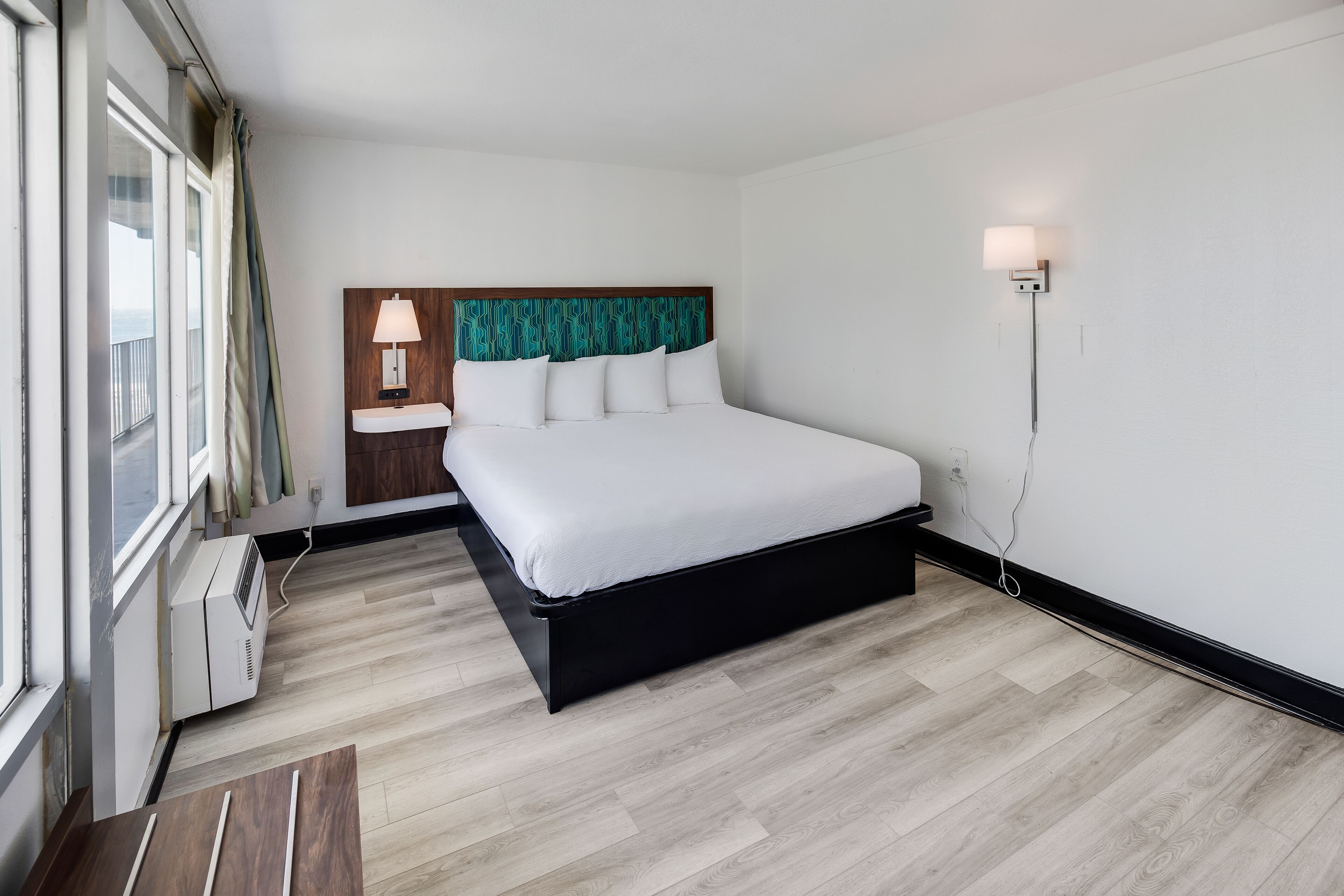 Ein modernes Hotelzimmer mit einem großen Bett, einem Nachttisch, einer Lampe und Blick ins Freie durch das Fenster.