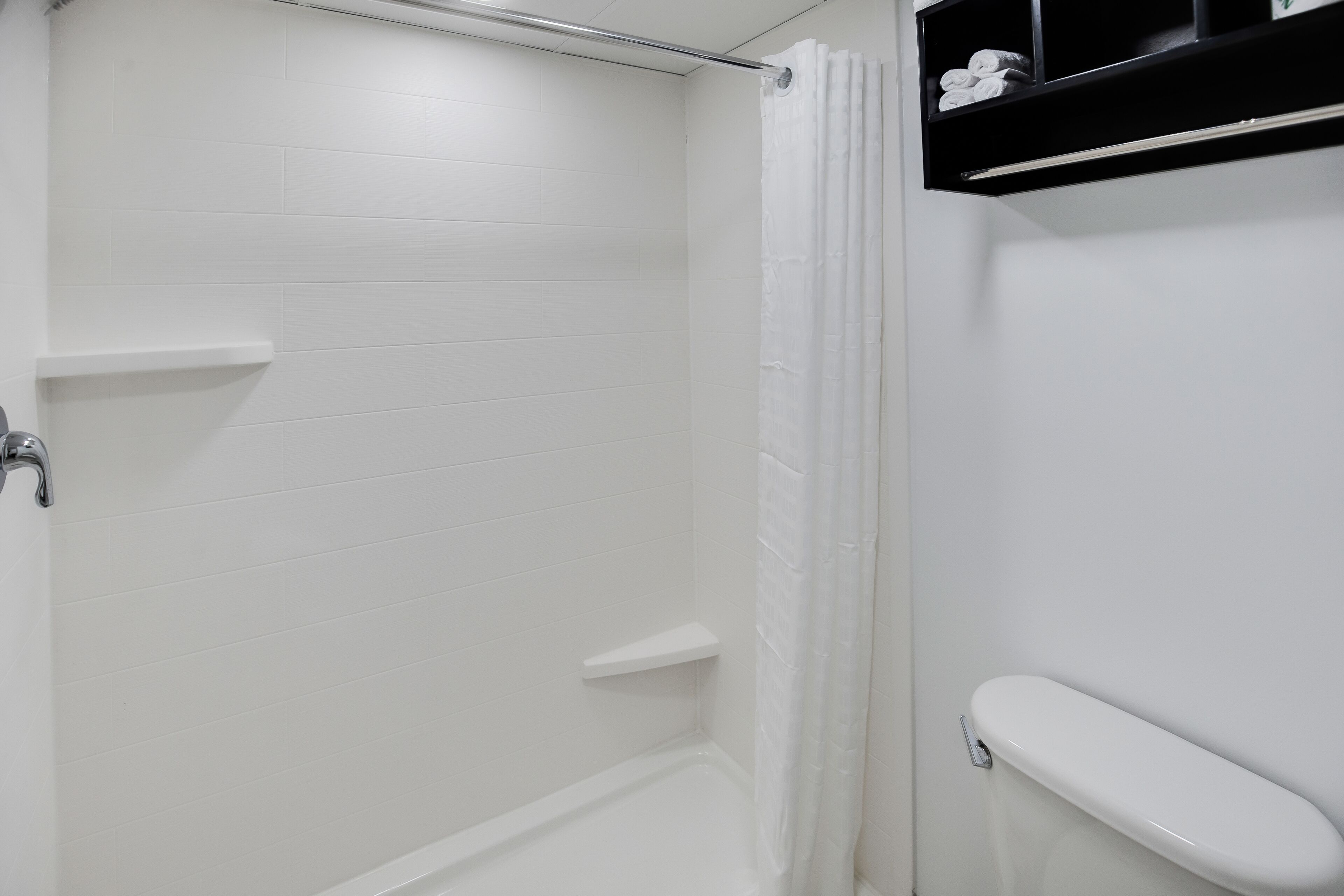 Un baño con paredes de azulejos blancos, una ducha, un inodoro y un estante con artículos de aseo.