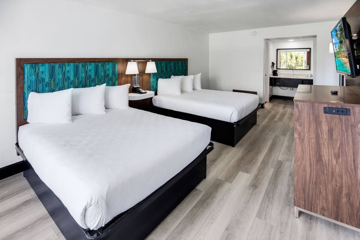 A hotel room with two beds, a wooden headboard, a desk with a TV, and a bathroom visible through an open door.