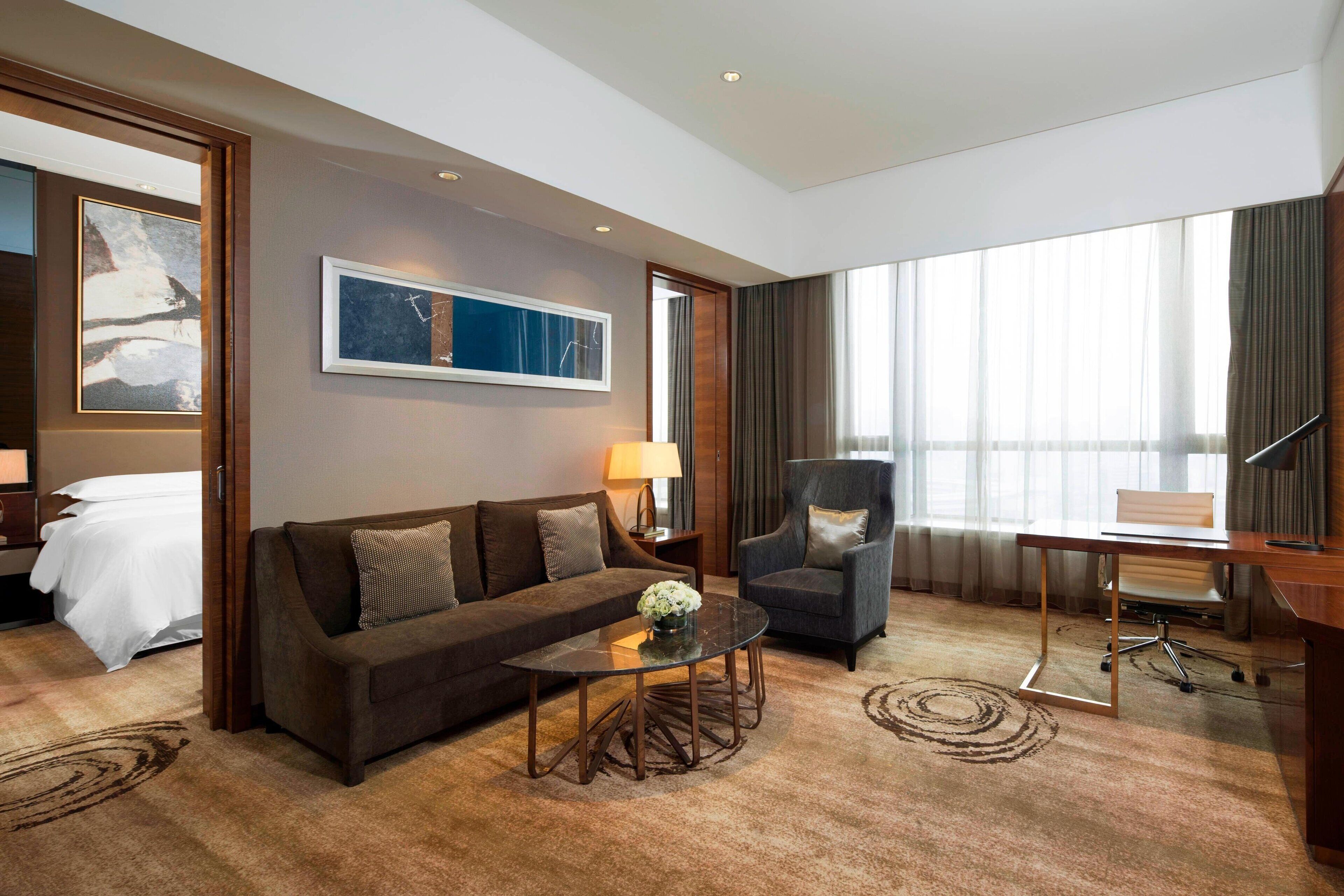 A hotel room with a sofa, armchair, desk, and a large window with curtains.