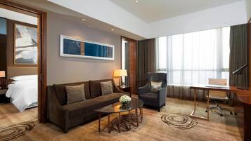 A hotel room with a sofa, armchair, desk, and a large window with curtains.