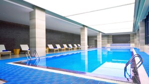 Una piscina interior con agua azul cristalina, rodeada de sillones de descanso y un suelo de baldosas.