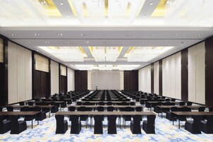 Un salón de conferencias con filas de mesas y sillas, una pantalla grande al frente y una alfombra estampada.