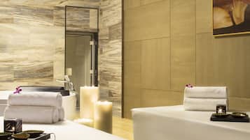 Relax in a serene spa room with a large mirror, bathtub, and two treatment tables, each equipped with towels, personal care items, and a candle.