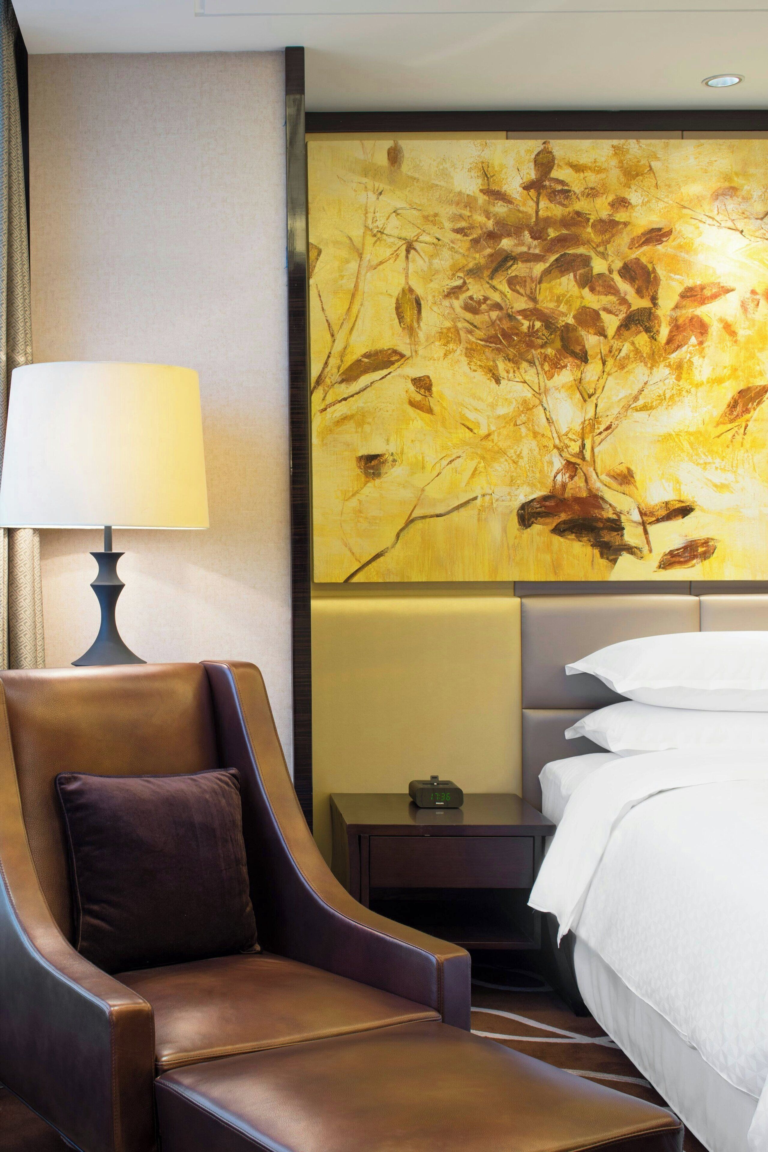 Ein Hotelzimmer mit braunem Ledersessel, einem Nachttisch mit Lampe, einem Bett mit weißer Bettwäsche und einem großen, abstrakten Gemälde an der Wand.