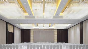 A conference room with rows of white chairs, a large screen at the front, and a ceiling with chandeliers.