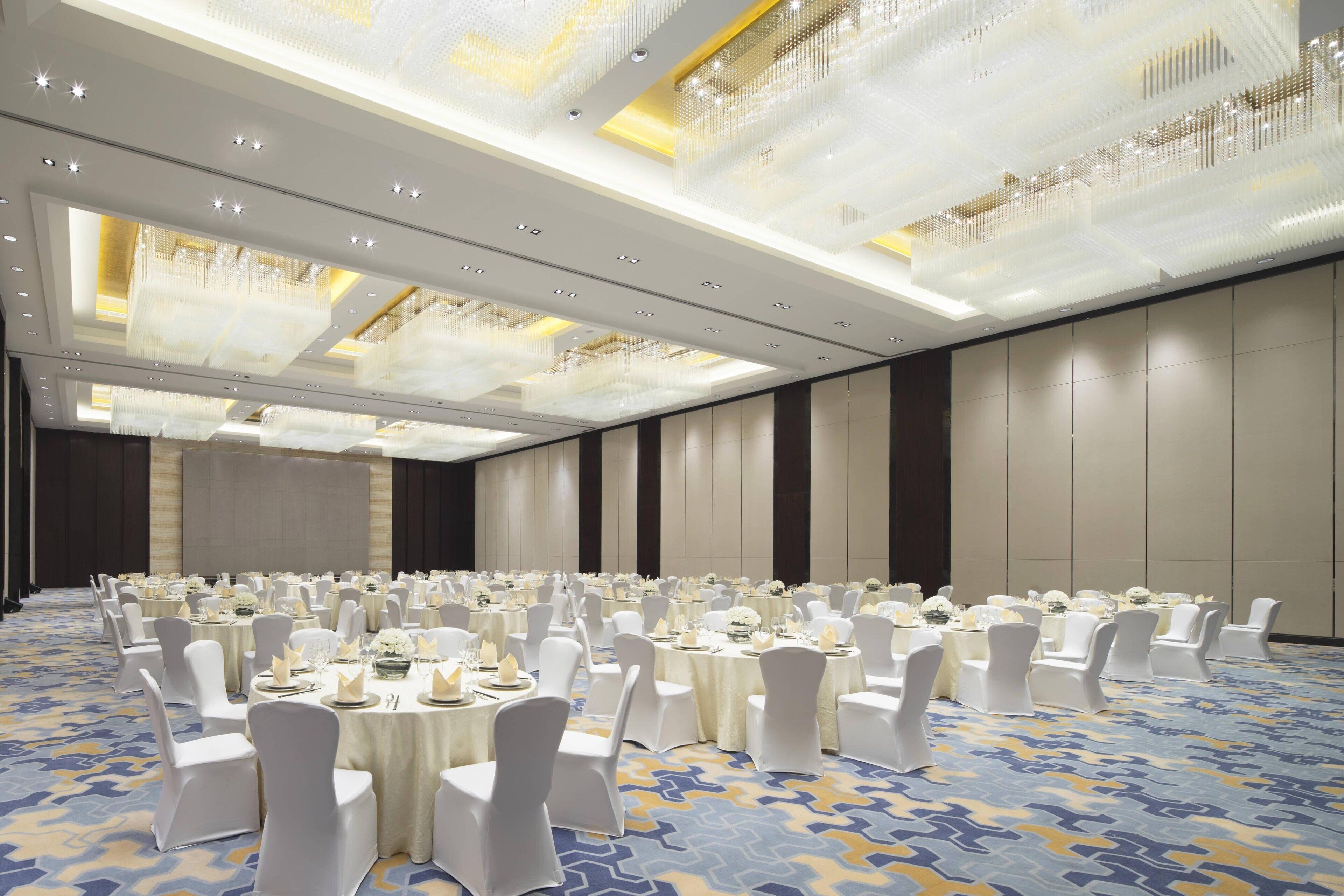 A large banquet hall with round tables set for events, featuring a patterned carpet and modern lighting fixtures.