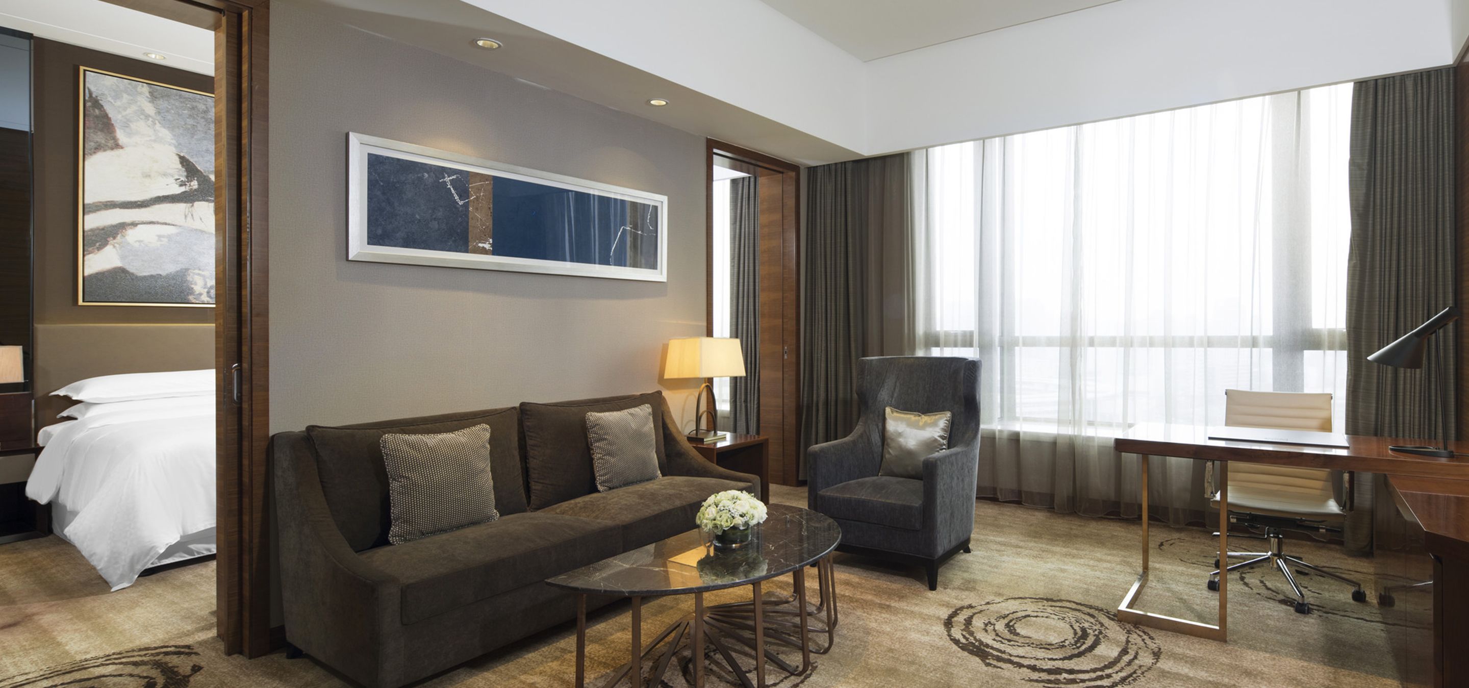 A hotel room with a sofa, armchair, desk, and a large window with curtains.